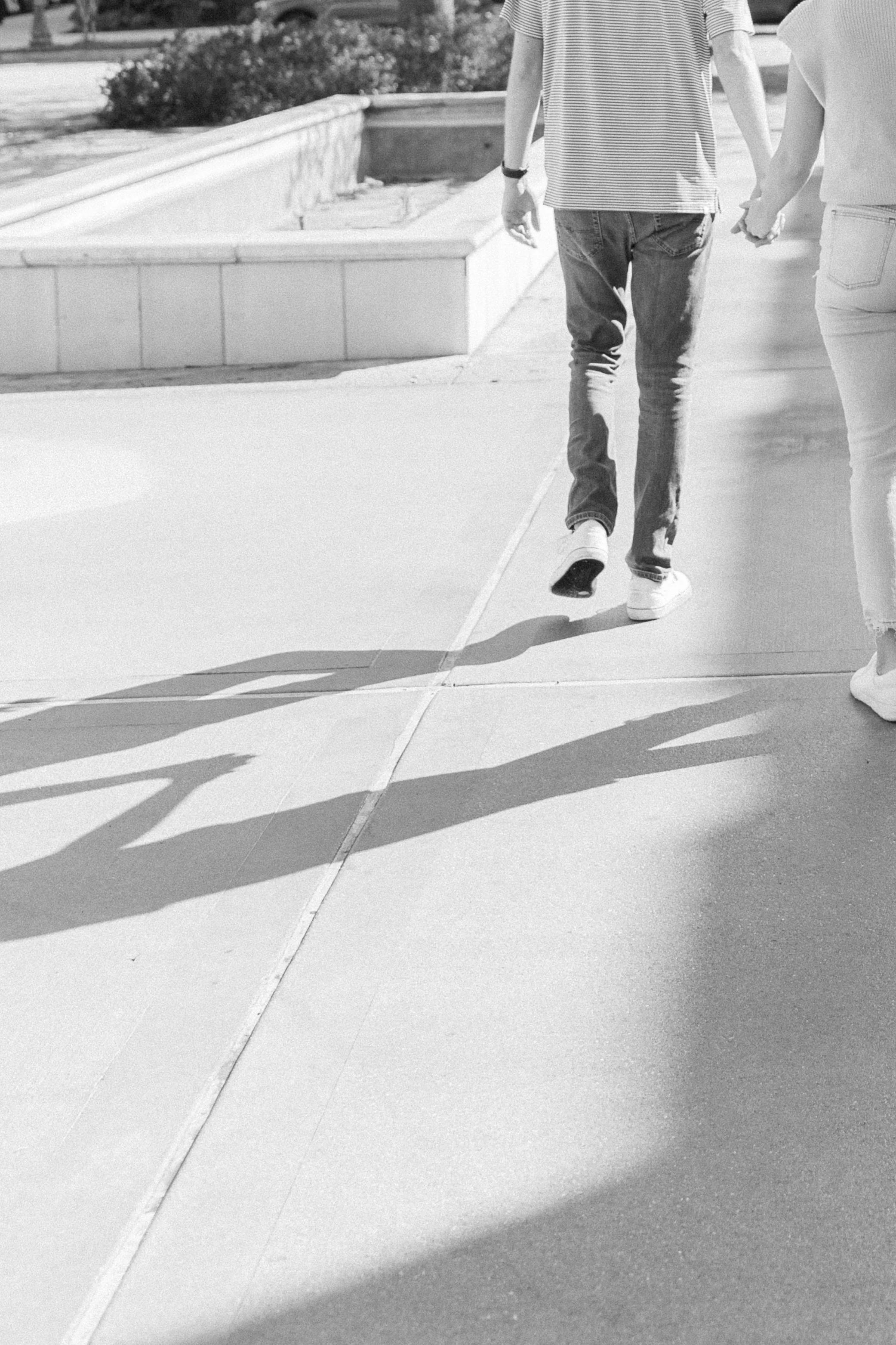 Two people holding hands walk on a sidewalk, their shadows cast on the ground.