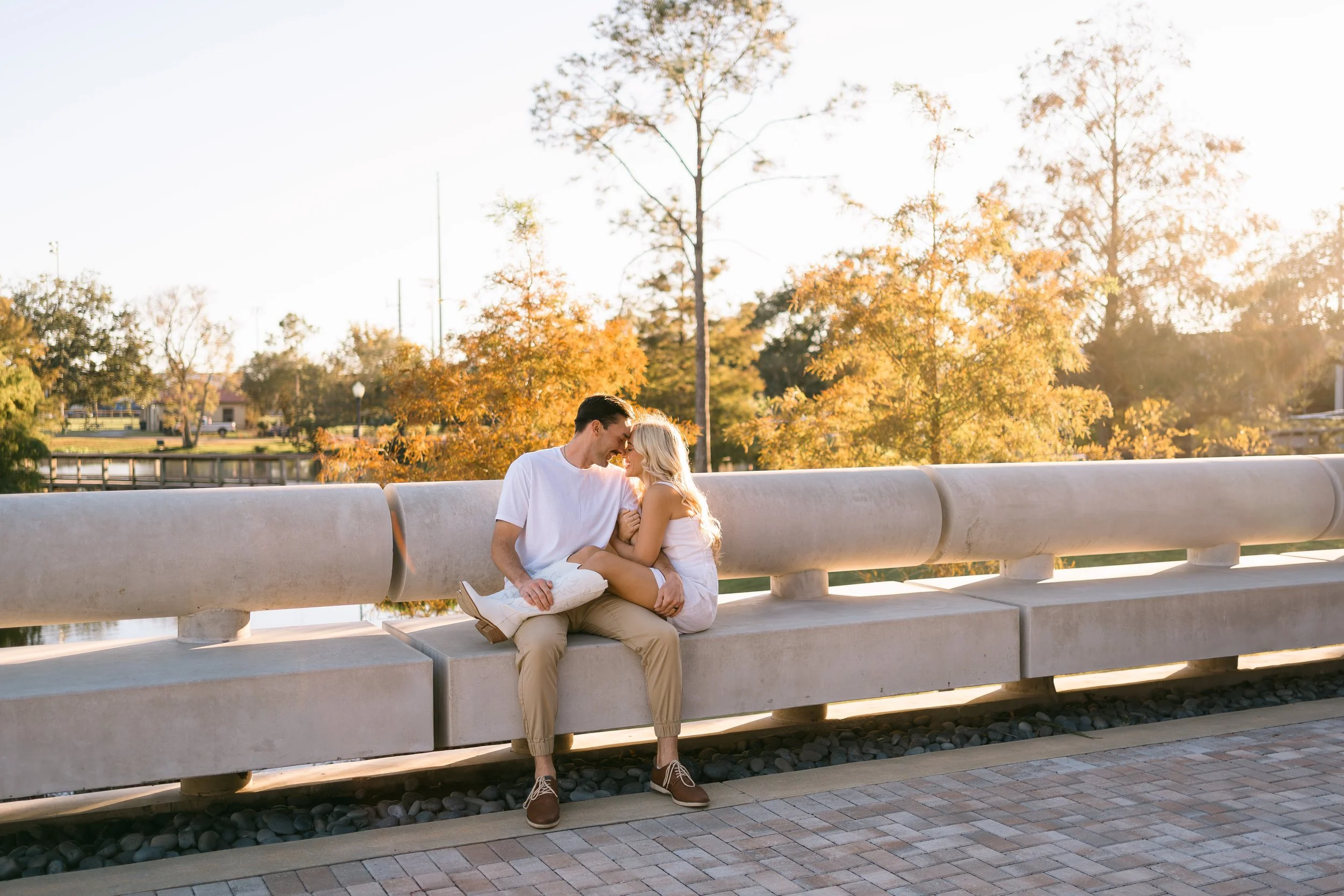 A young couple sits on a concrete bench outdoors during golden hour. They sit close, touching foreheads, smiling, with autumn trees in the background.