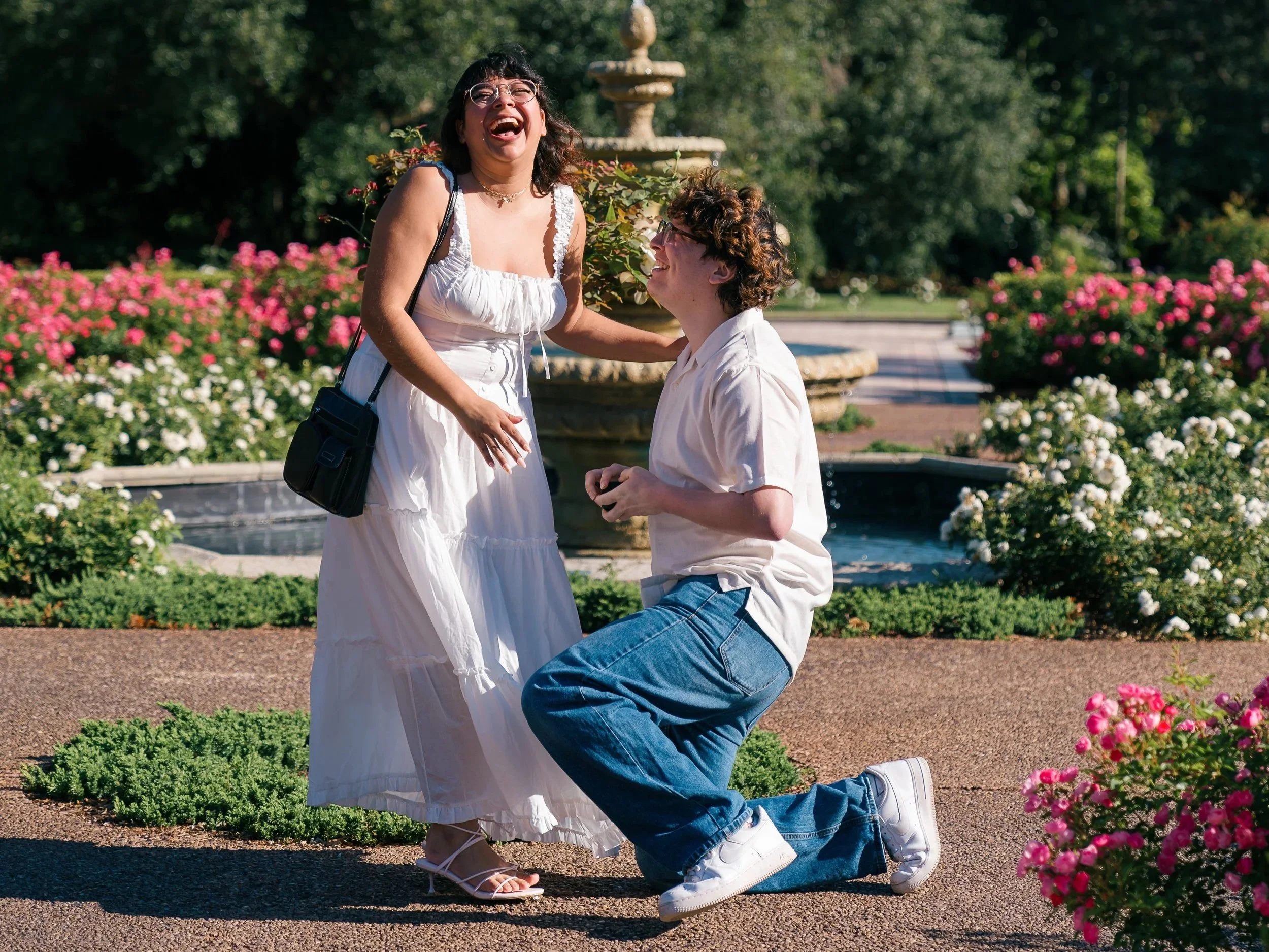A woman wearing a white dress is laughing while a man in a white shirt and jeans kneels in front of her, holding her hand. They are outdoors in a garden with colorful flowers and greenery.