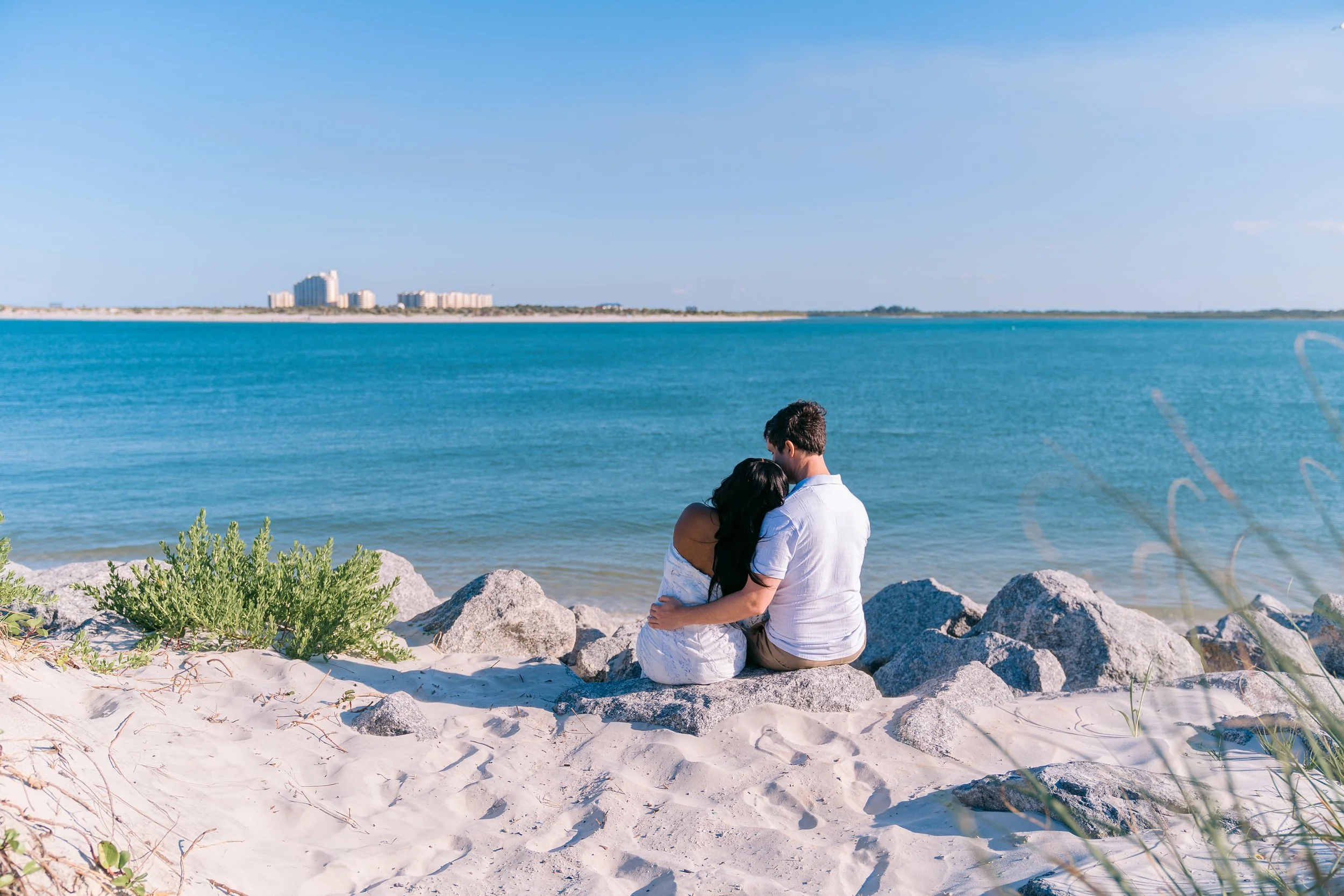 A couple sitting on rocks by the beach, embracing, with a city skyline across the water in the background.
