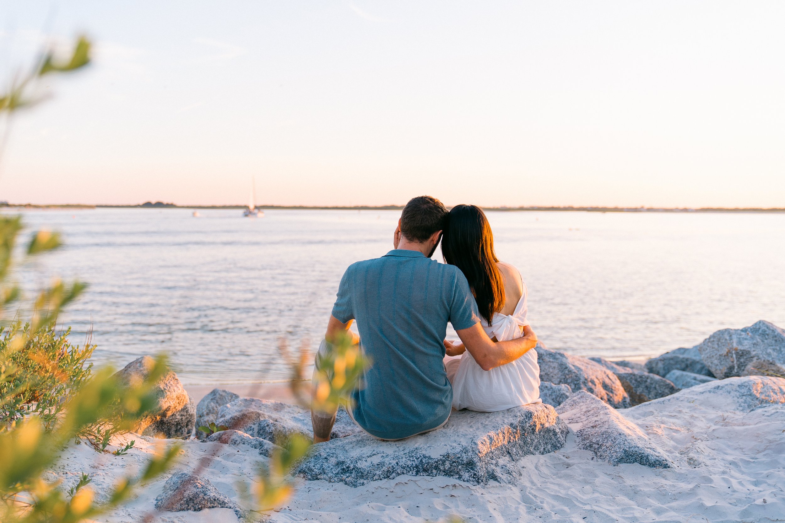 A couple sitting on rocks by the water during sunset, with a sailboat in the distance.