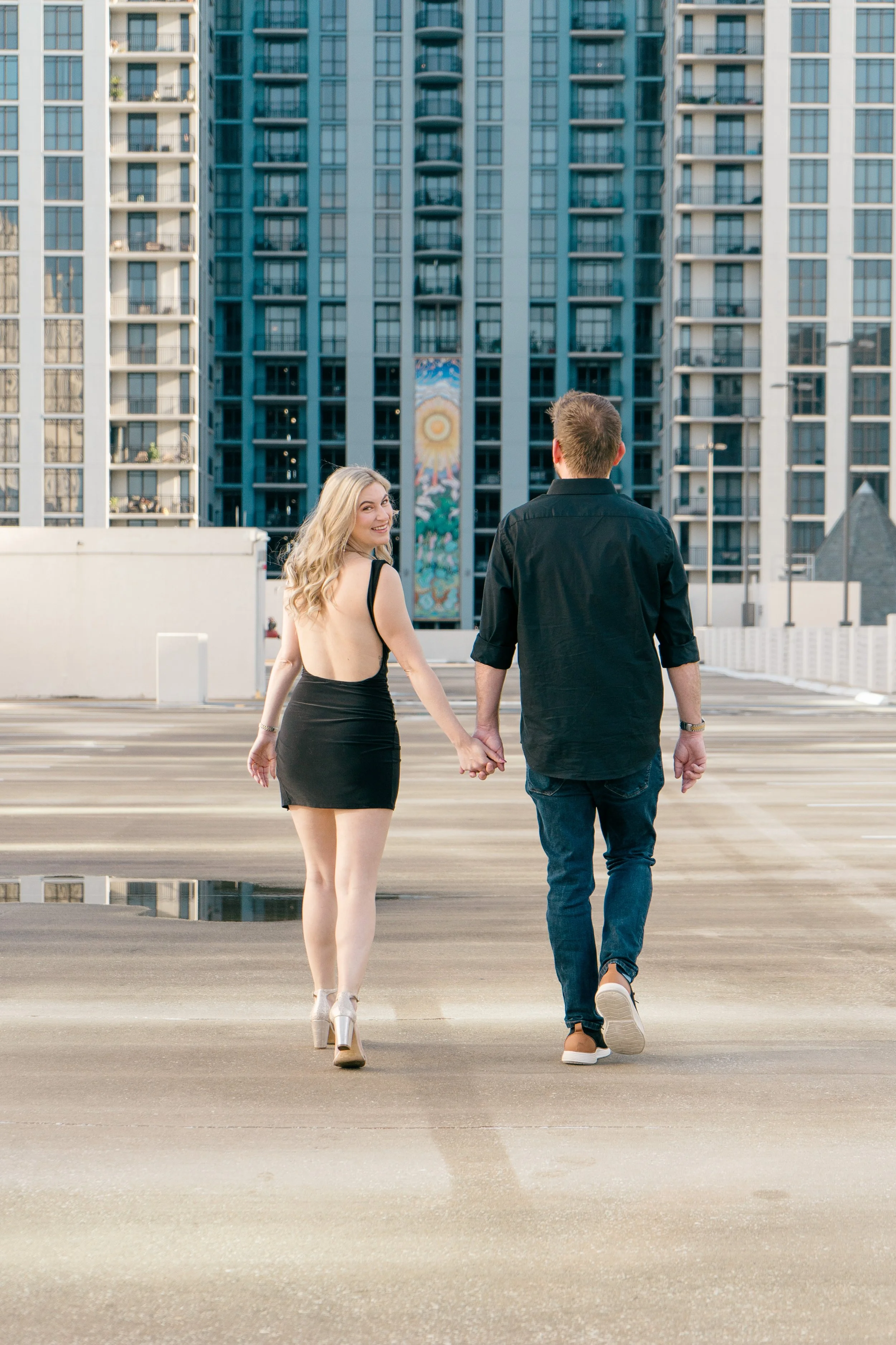 A couple holding hands and walking away in an urban parking lot, with tall apartment buildings in the background.