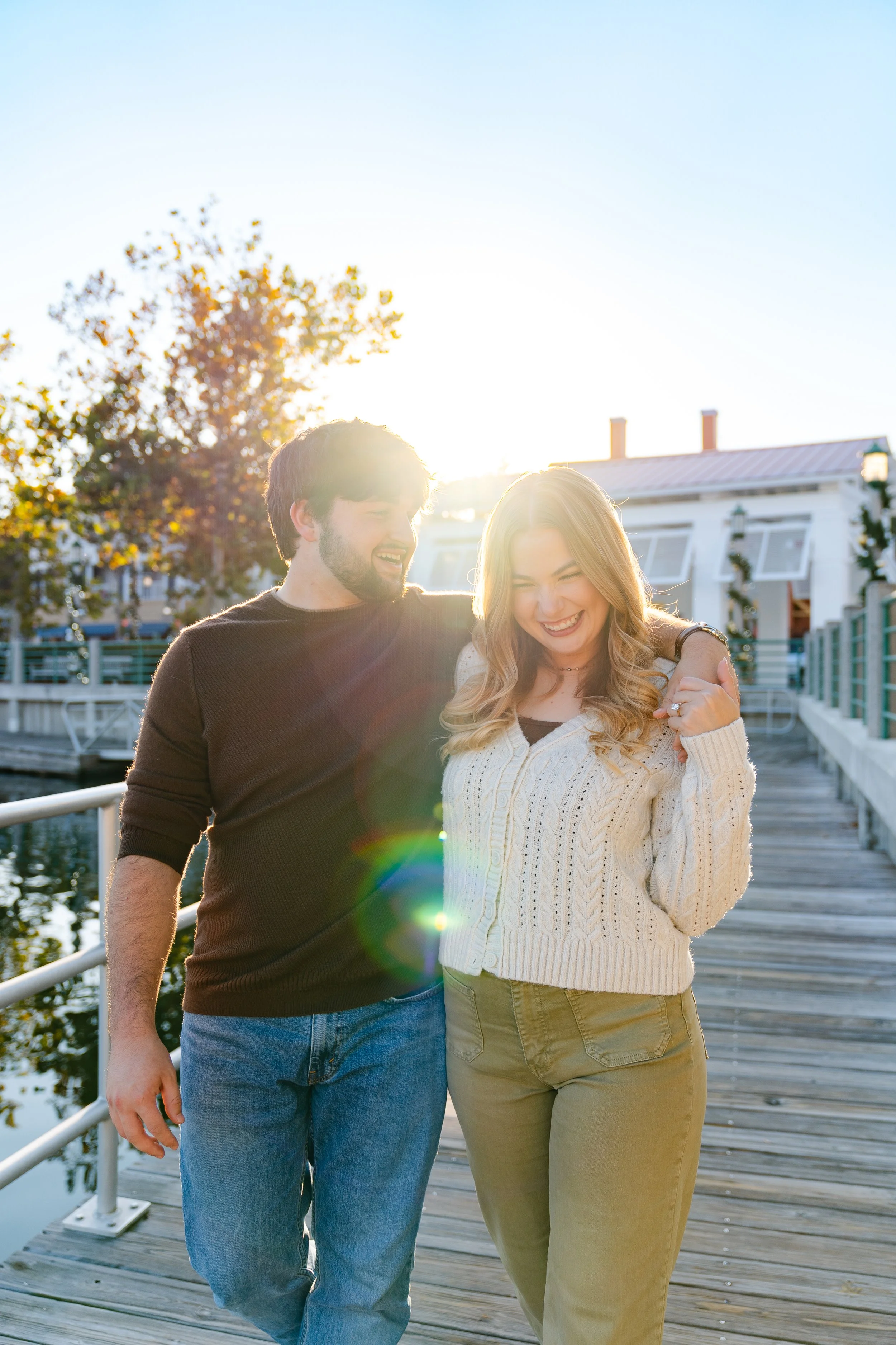 A smiling man and woman walk arm in arm on a wooden dock by the water in the late afternoon sun.