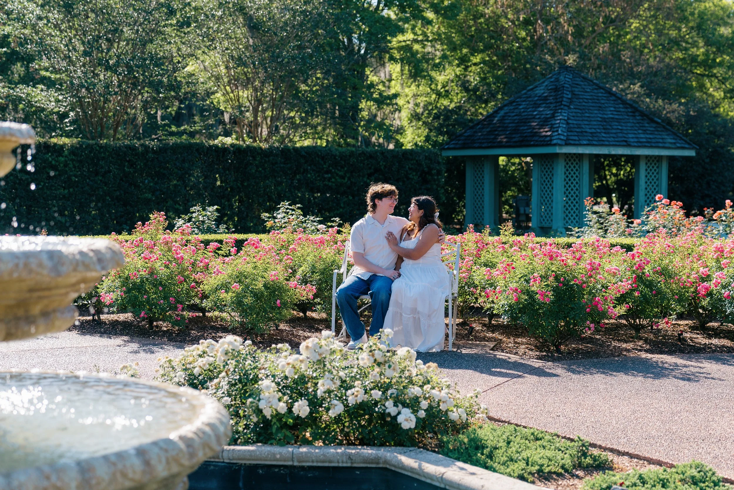 A couple sitting on a white bench in a garden with pink and white roses, surrounded by greenery, with a gazebo in the background and a fountain in the foreground.