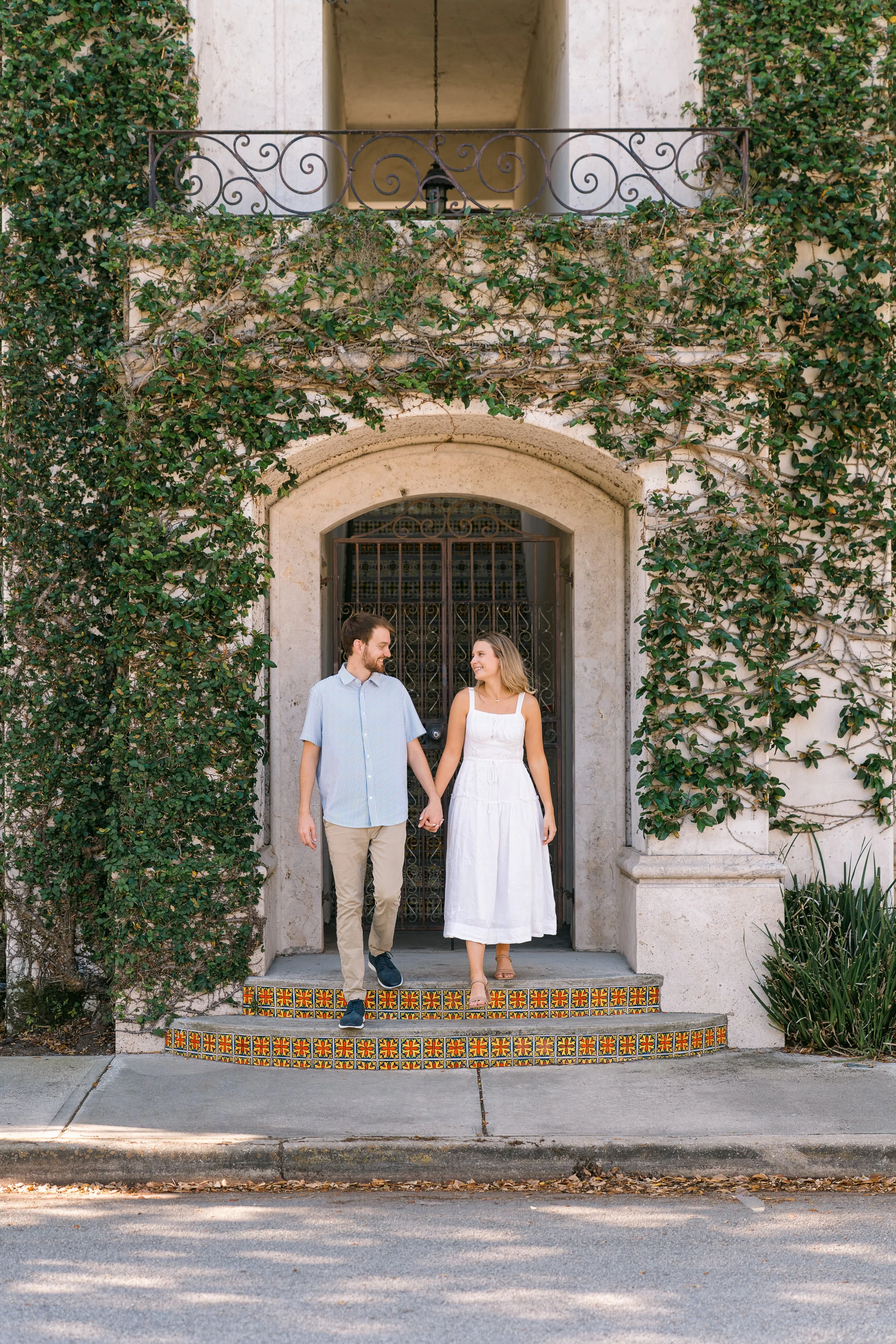 A young couple holding hands and walking out of a doorway decorated with colorful tiles, surrounded by ivy-covered walls.