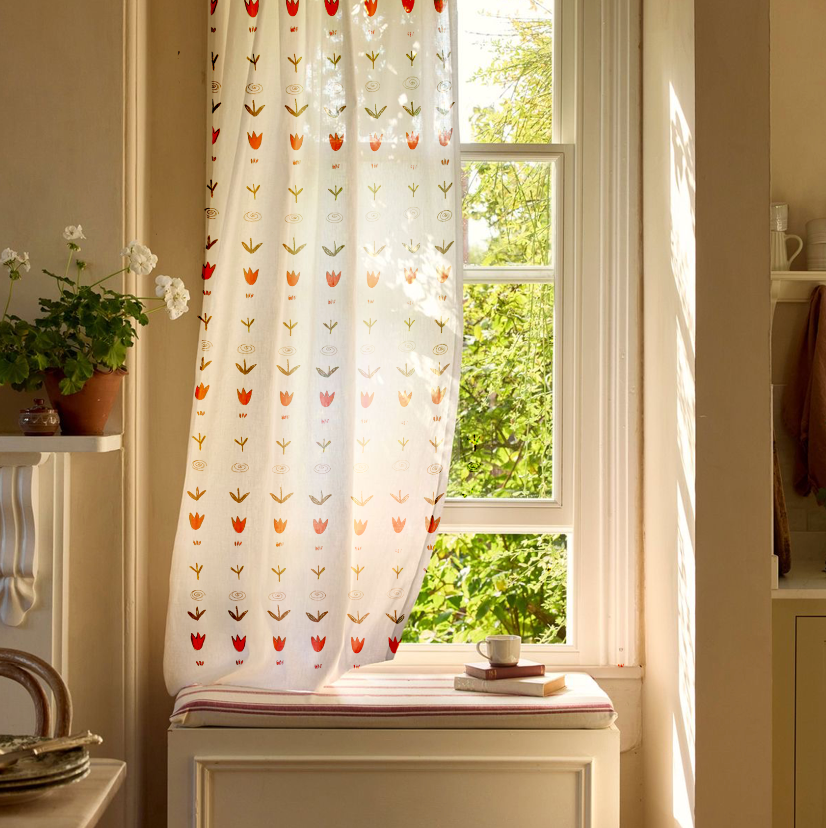 Sunlight coming through a window with a white curtain decorated with colorful tulip patterns, a cushioned window seat with a mug and books, and greenery outside.