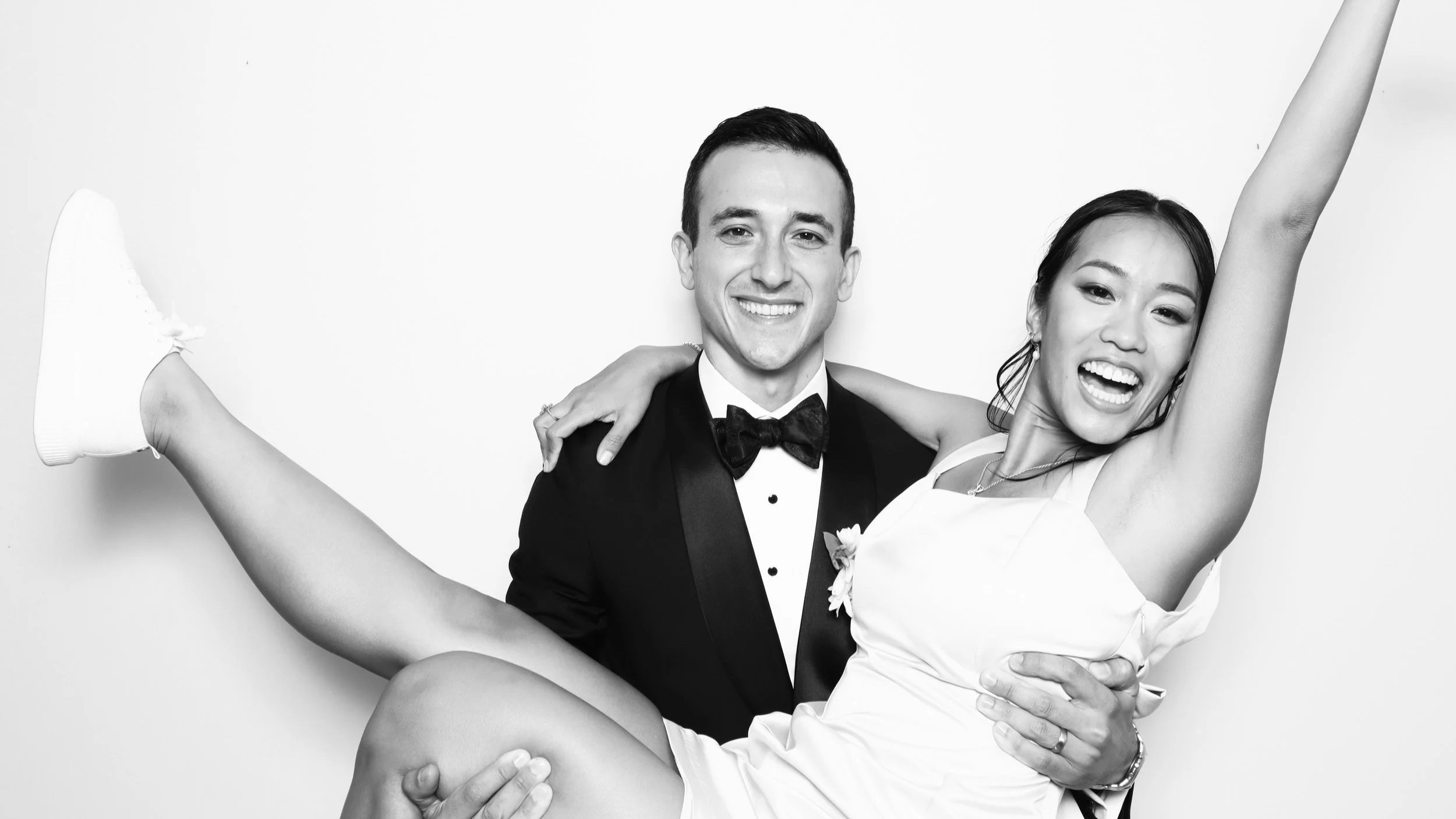 A smiling man dressed in a tuxedo holding a laughing woman in a white dress. The woman is raising one arm and has her legs extended outwards.