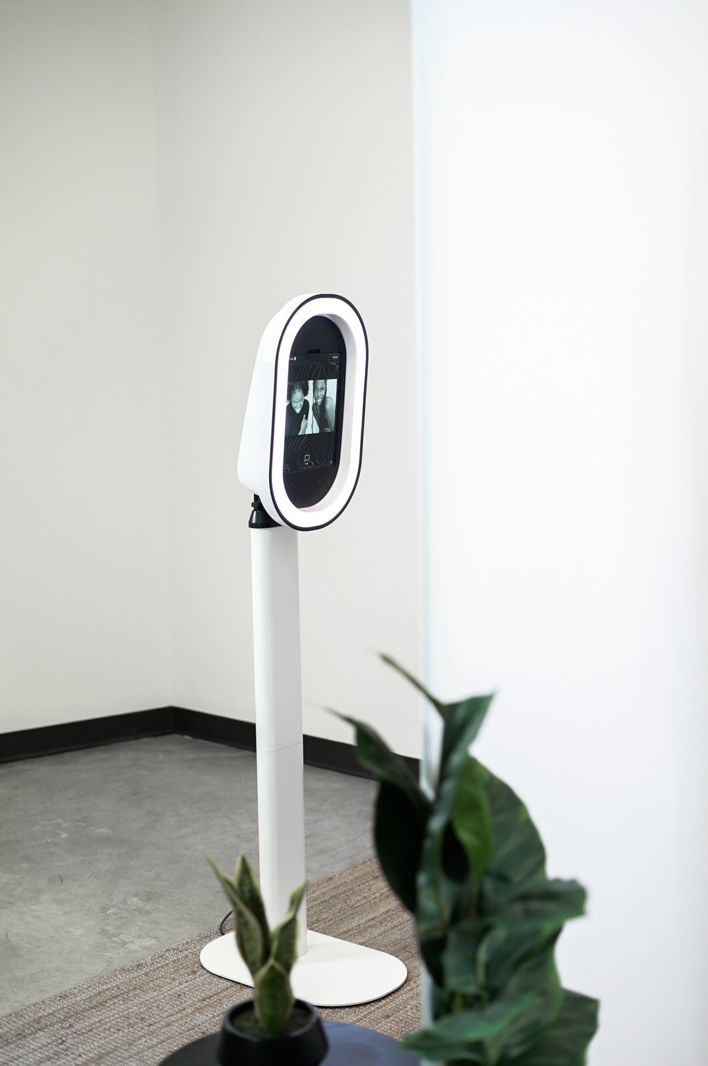 A digital kiosk with a screen showing black-and-white photos of two people, situated in a minimalistic room with white walls, a gray floor, and a potted plant in the foreground.