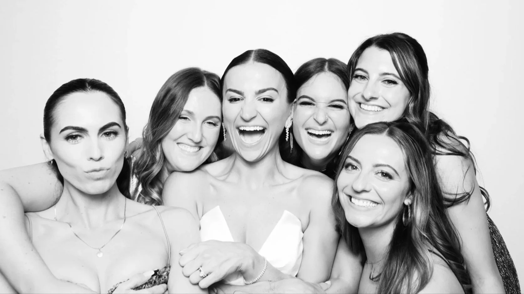 Group of six women smiling and laughing together, posing for a black and white photo, with one woman at the center wearing a strapless dress and earrings.