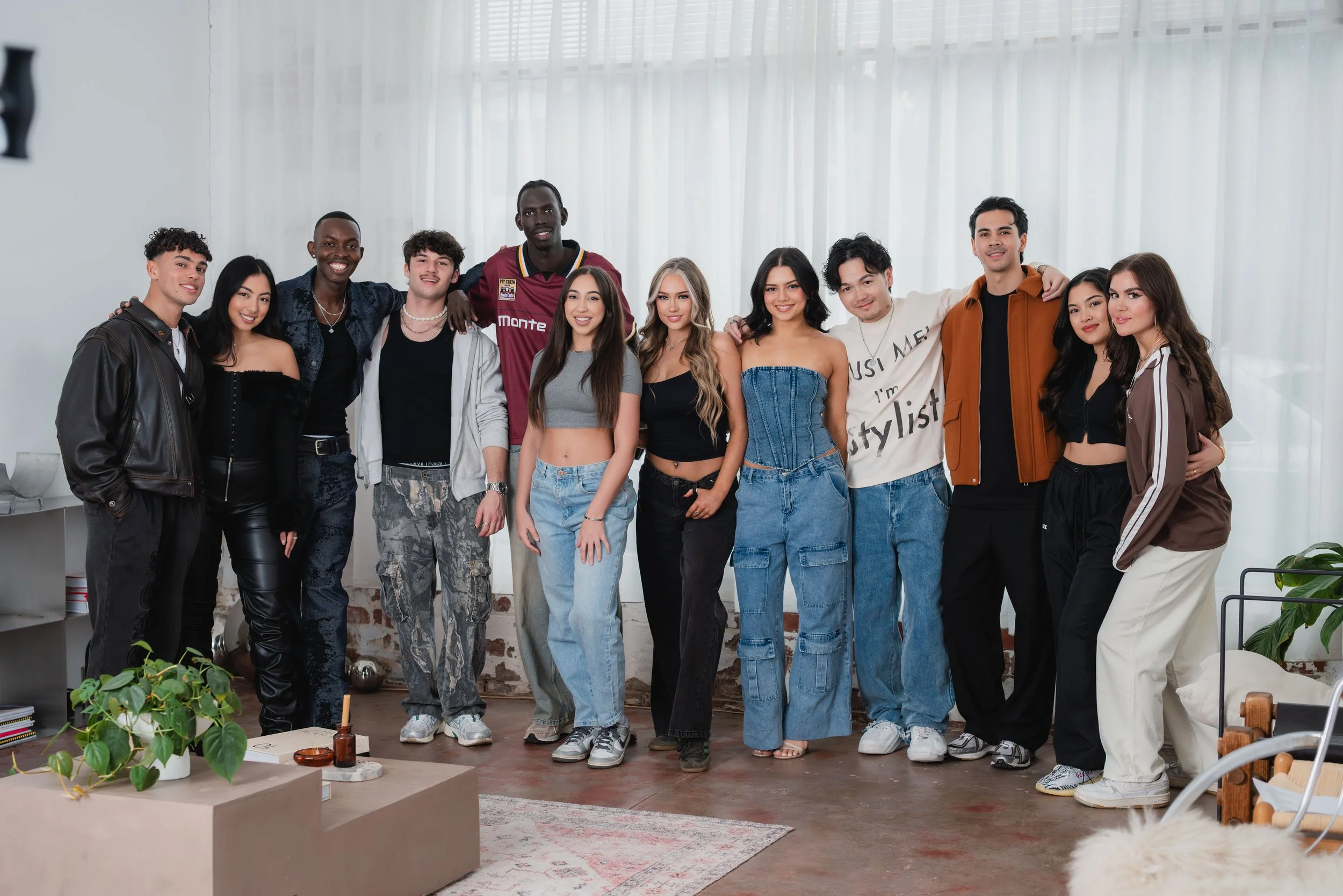 Group of eleven young adults standing together in a well-lit room with white curtains and plants, smiling at the camera.