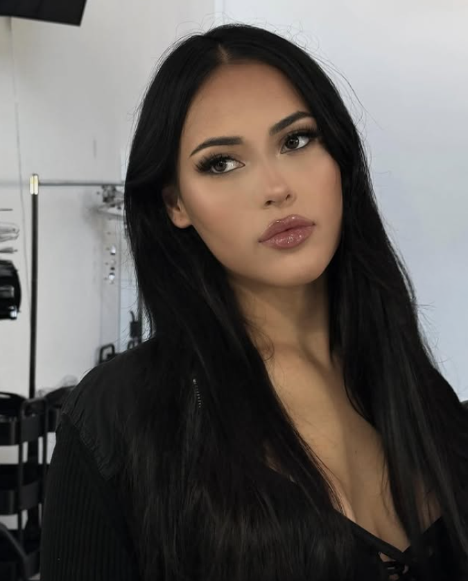 A young woman with long black hair, makeup, and glossy lips, looking slightly to the side in a well-lit indoor setting.