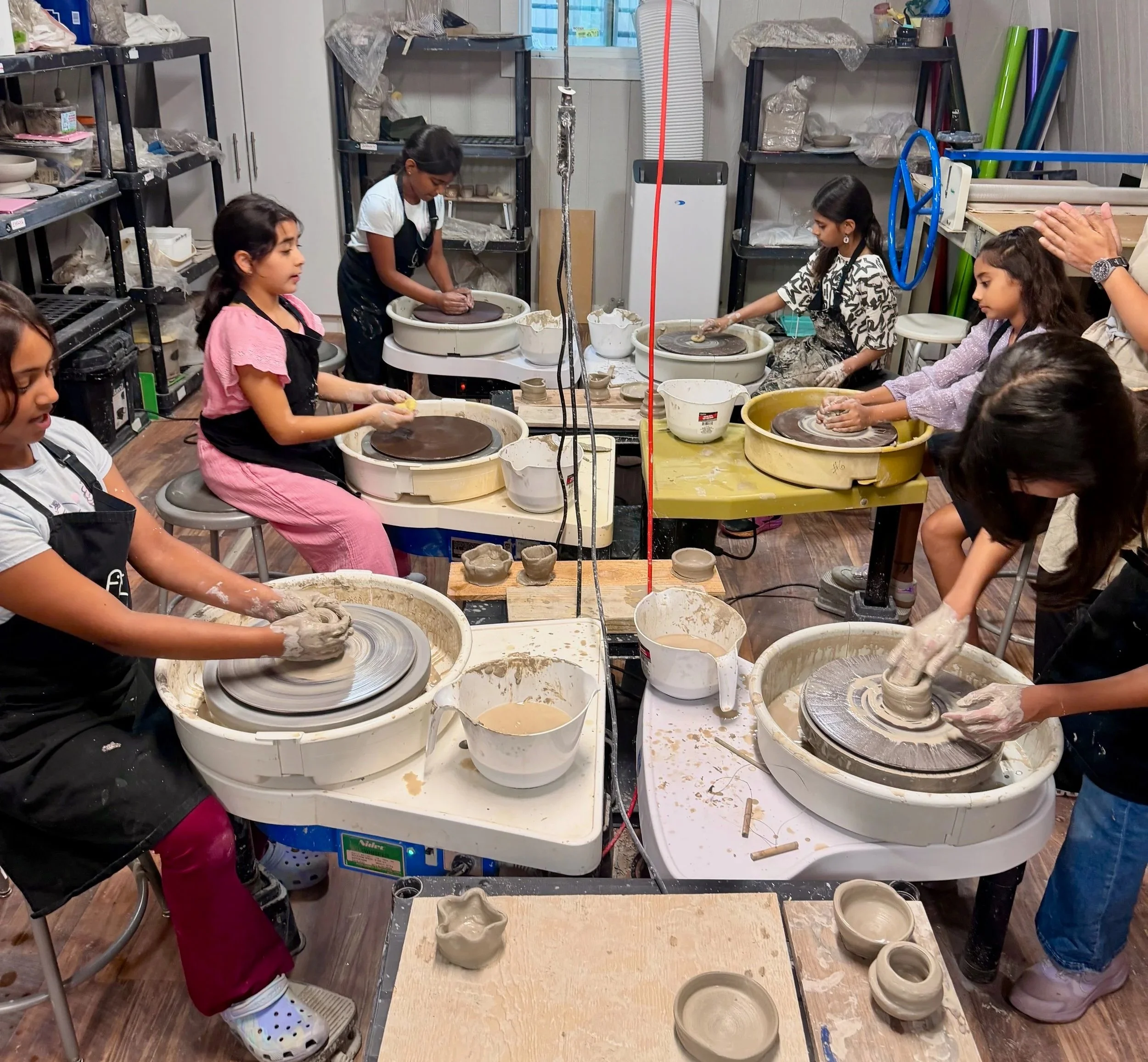 Children and adults participating in a ceramics class, working on their pottery wheels in a studio filled with shelves of supplies and finished ceramic pieces.
