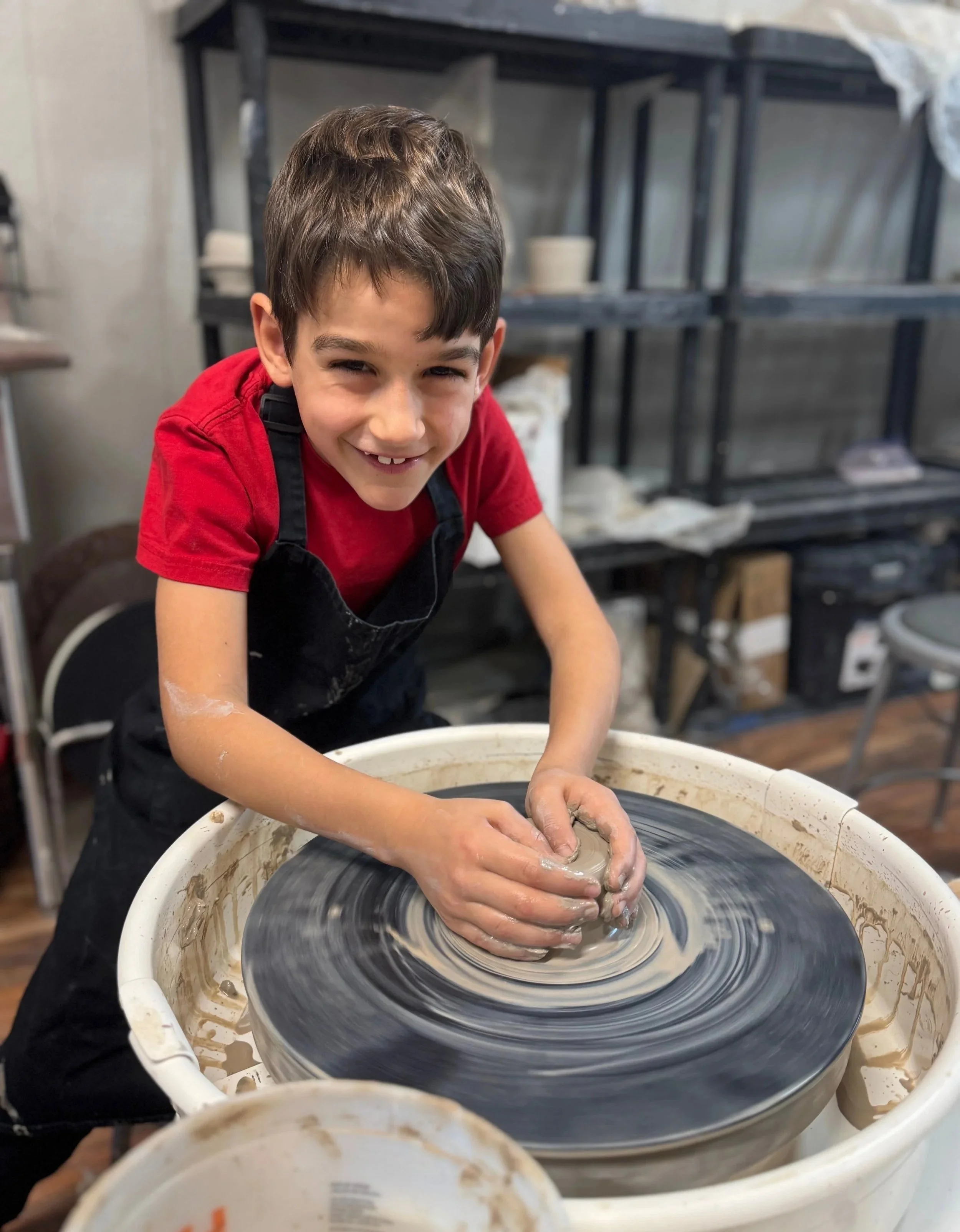 A young boy with a red shirt and black apron smiling while shaping clay on a pottery wheel in a ceramic studio.