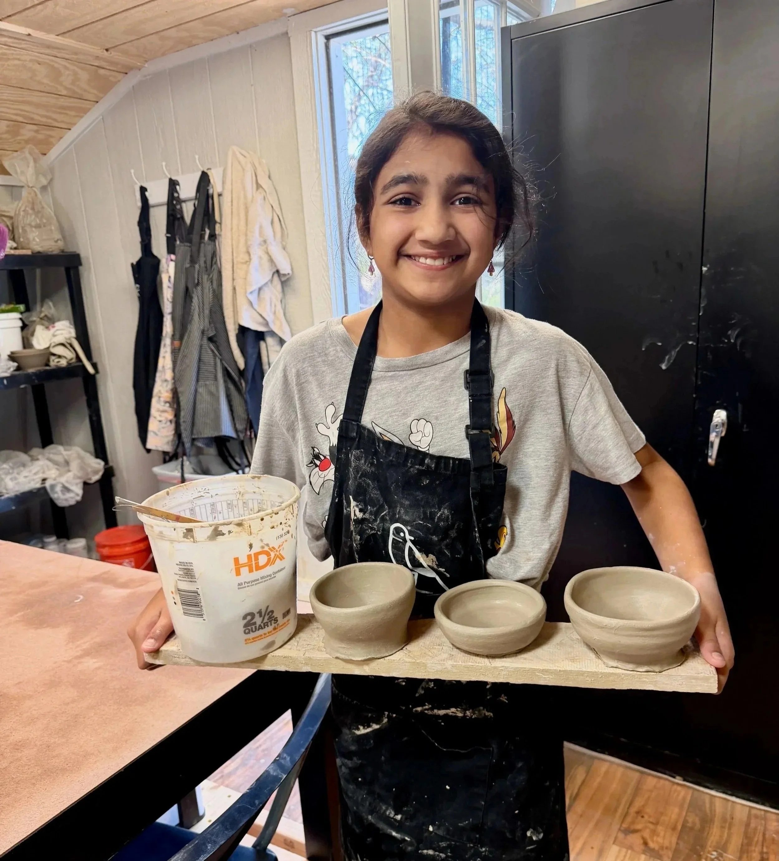 Young girl smiling and holding a wooden board with four small ceramic bowls and a container of clay for pottery.