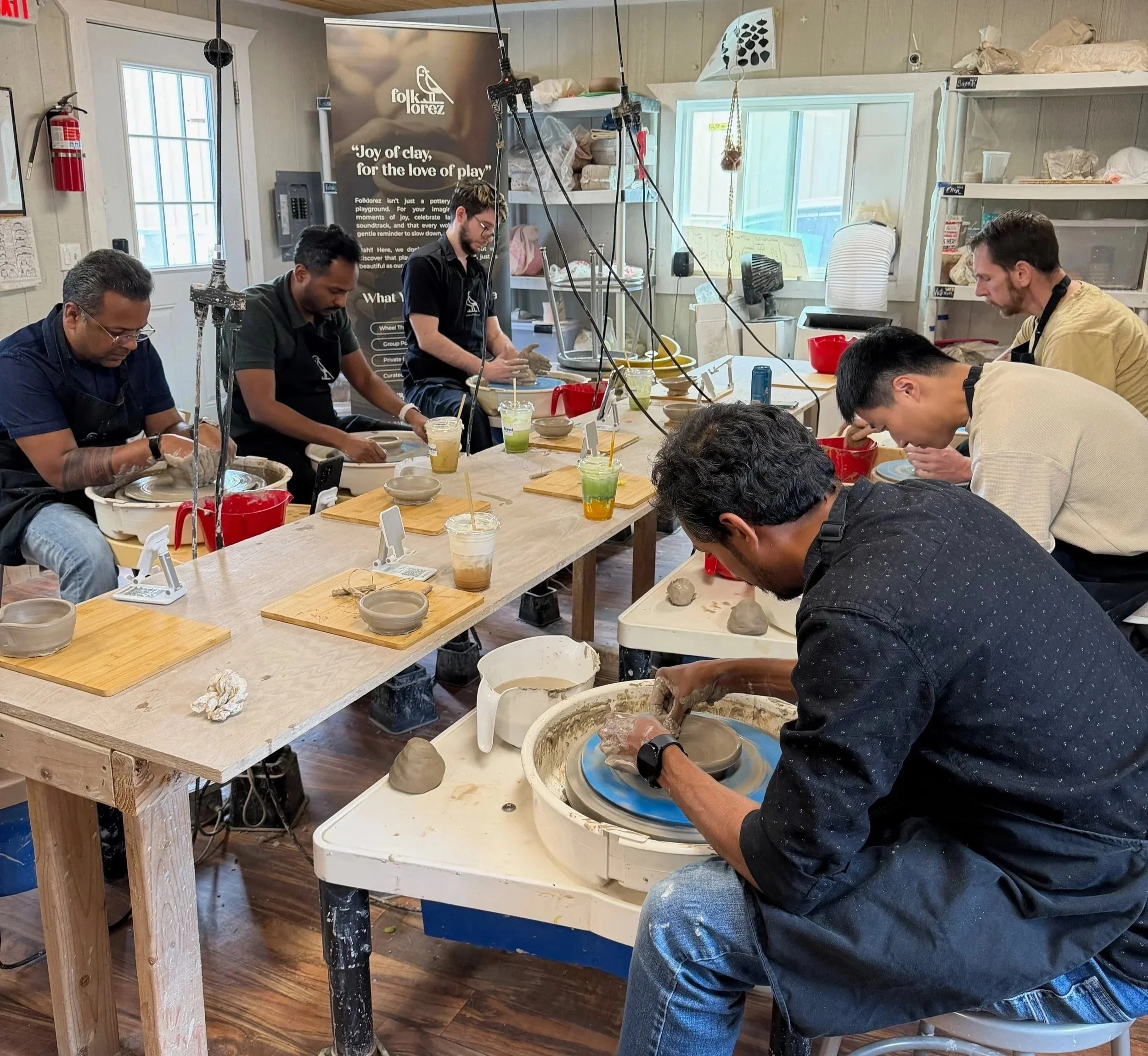 People participating in a pottery class working on clay pottery on spinning wheels in a well-lit studio.