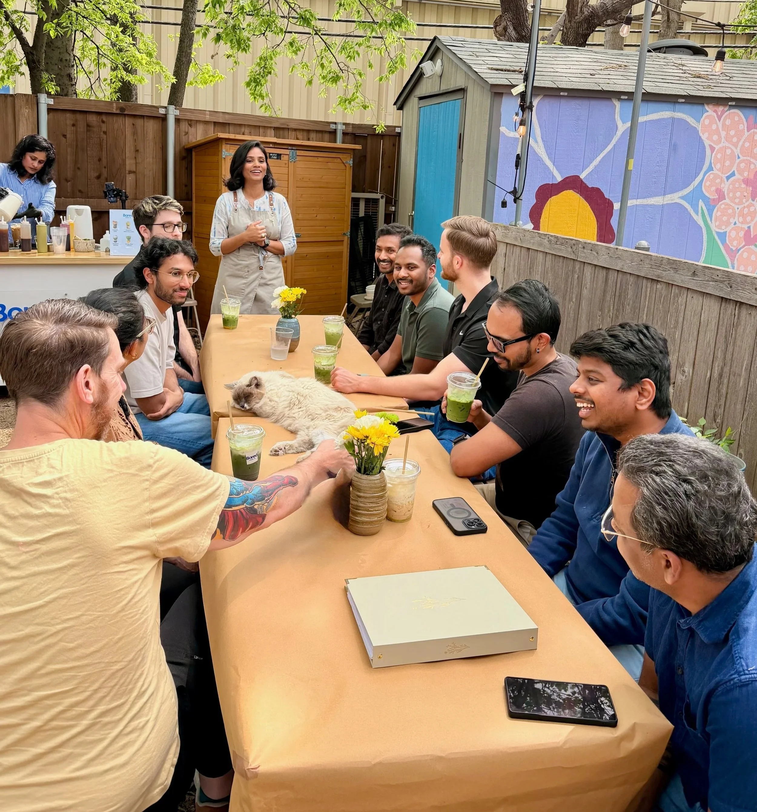 A group of people sit around a long outdoor table, smiling and engaging in conversation, with beverages and a cat on the table. A woman stands at the head of the table, speaking to the group. There are trees and colorful murals in the background.