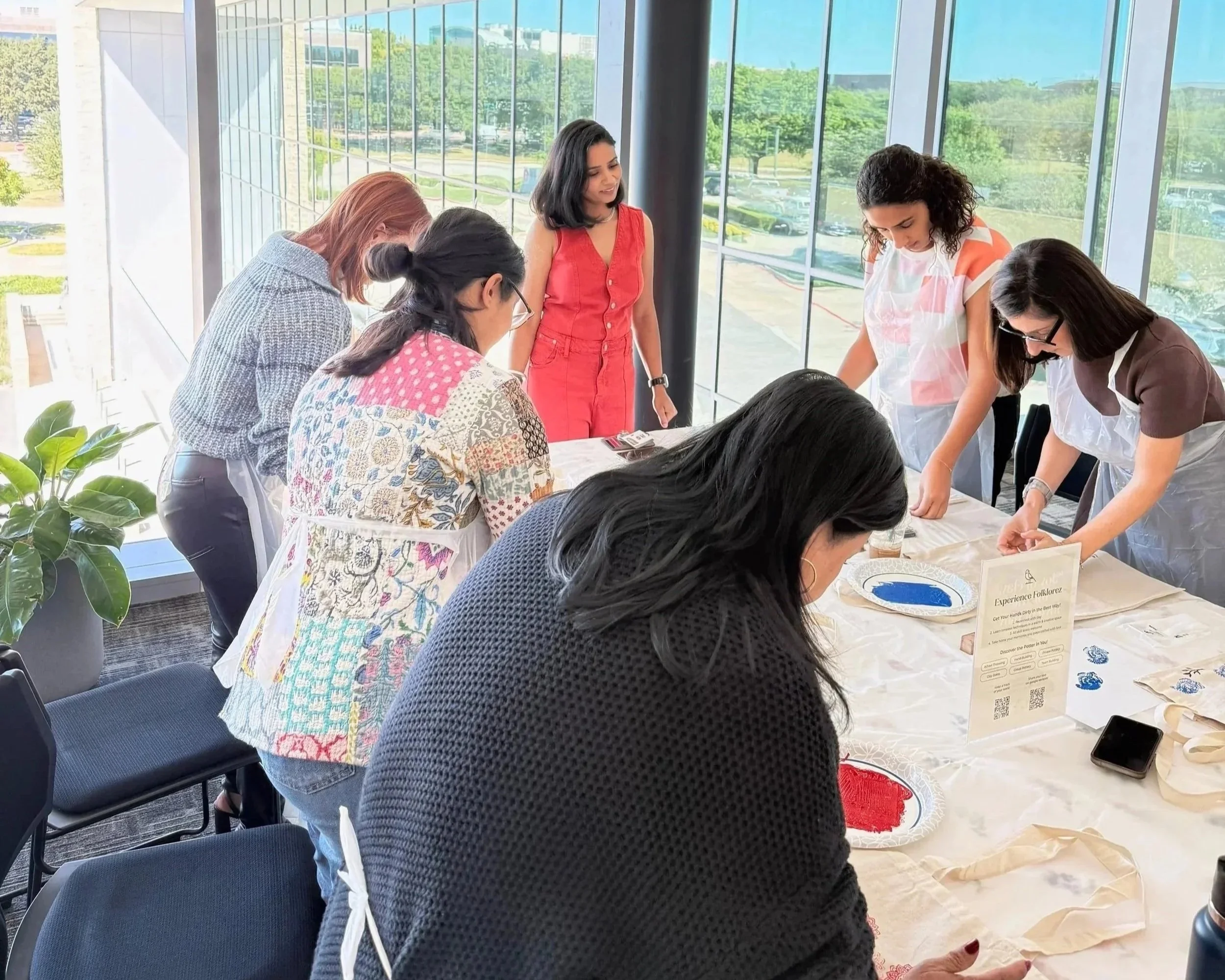 Women participating in a ceremonial activity in a bright room with large windows, some are bending over a table with colored powders, and others are standing, observing or assisting.