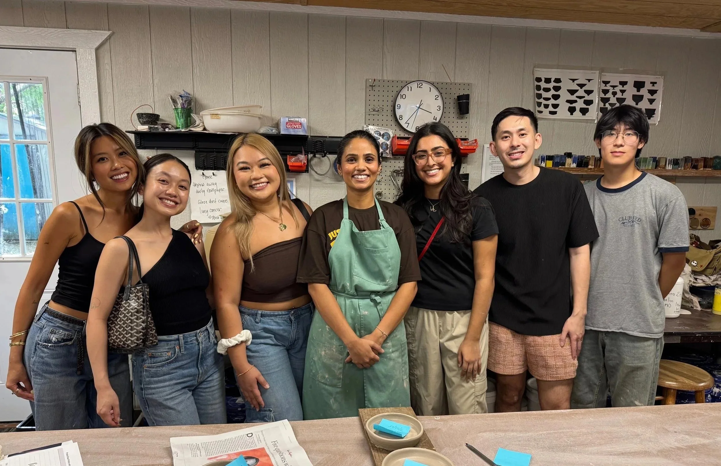 A group of eight people standing together indoors, smiling at the camera. They are in a workshop or kitchen with various tools and supplies on shelves behind them.