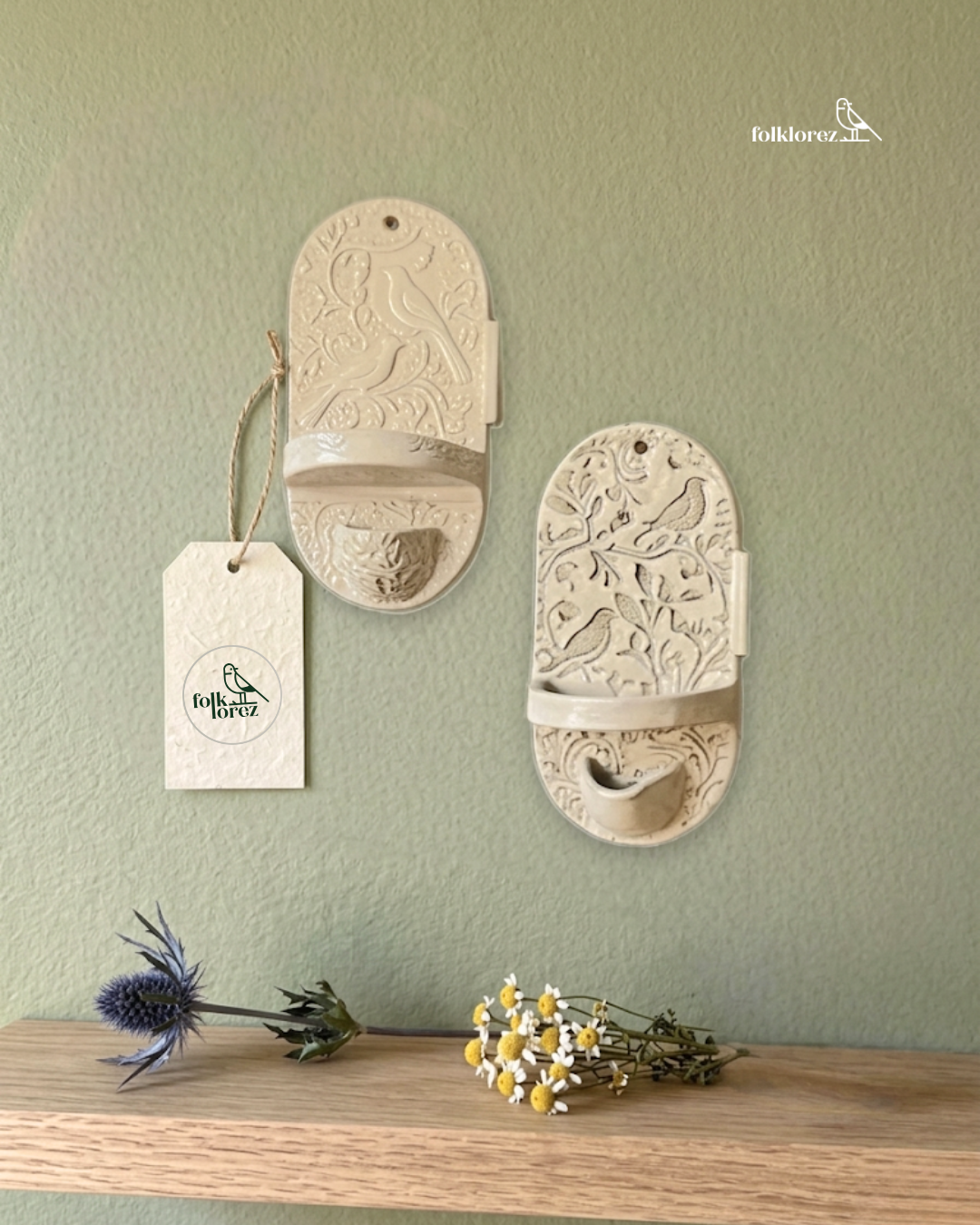 Two decorative wall planters with embossed bird and branch patterns hanging on a green wall, a small paper tag on the left planter labeled 'folklorez,' and a wooden surface below with a sprig of purple thistle and a small bunch of white and yellow daisies.