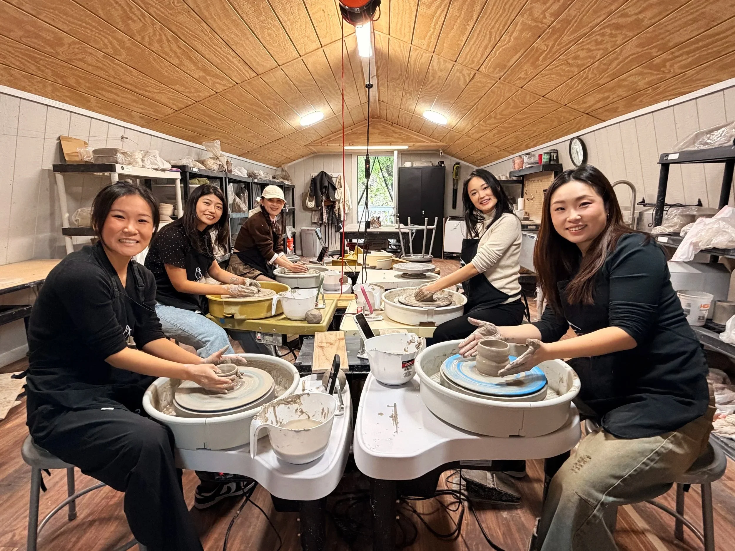 Six women participating in a pottery class, working on pottery wheels in a rustic studio with shelves full of supplies.
