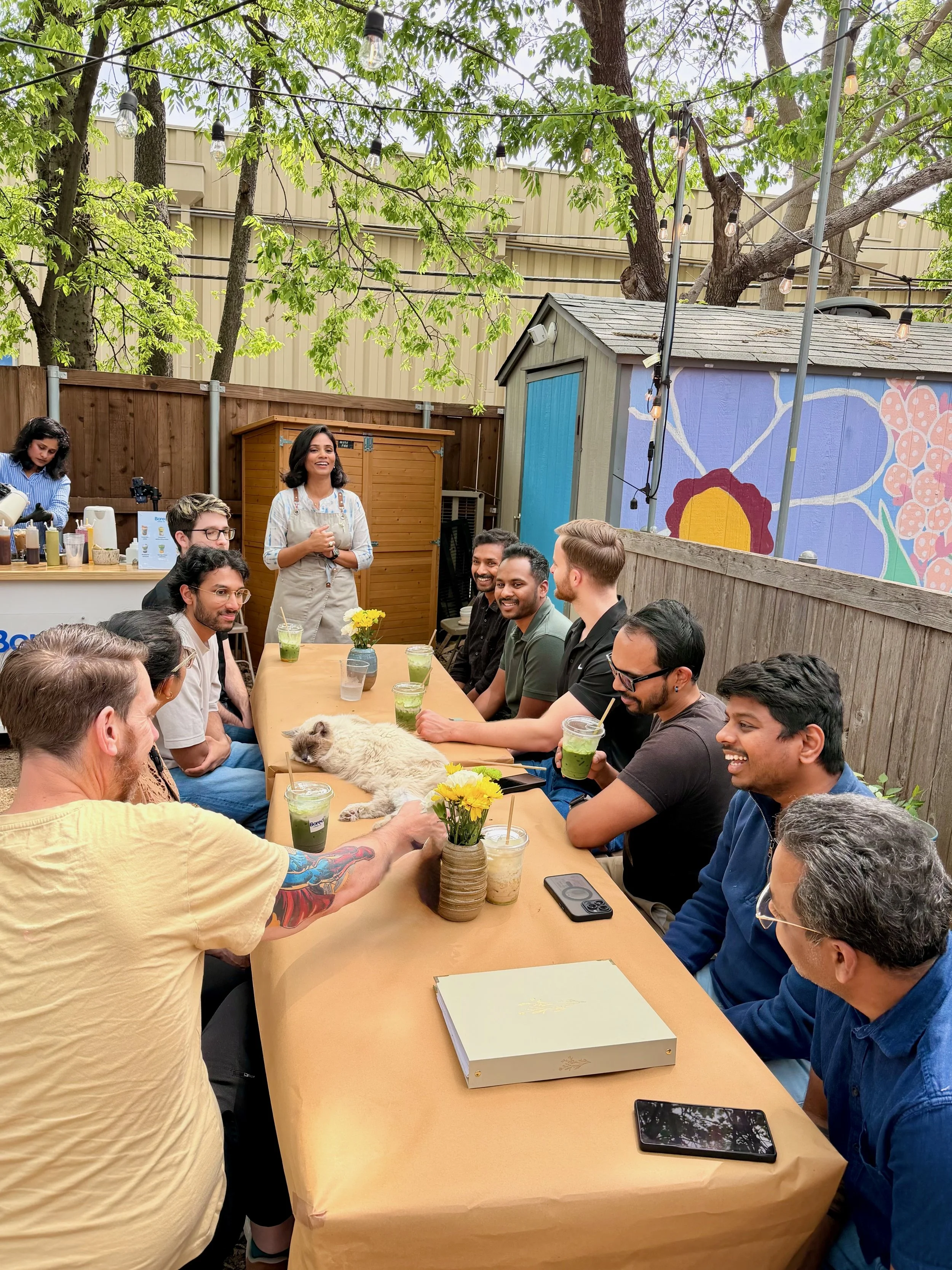 A group of people gathered around a table outdoors, having a celebration or meeting. There are drinks, flowers, a cat lying on the table, and a woman standing and speaking to the group. The setting is a backyard with string lights, trees, and a colorful mural on a small shed in the background.
