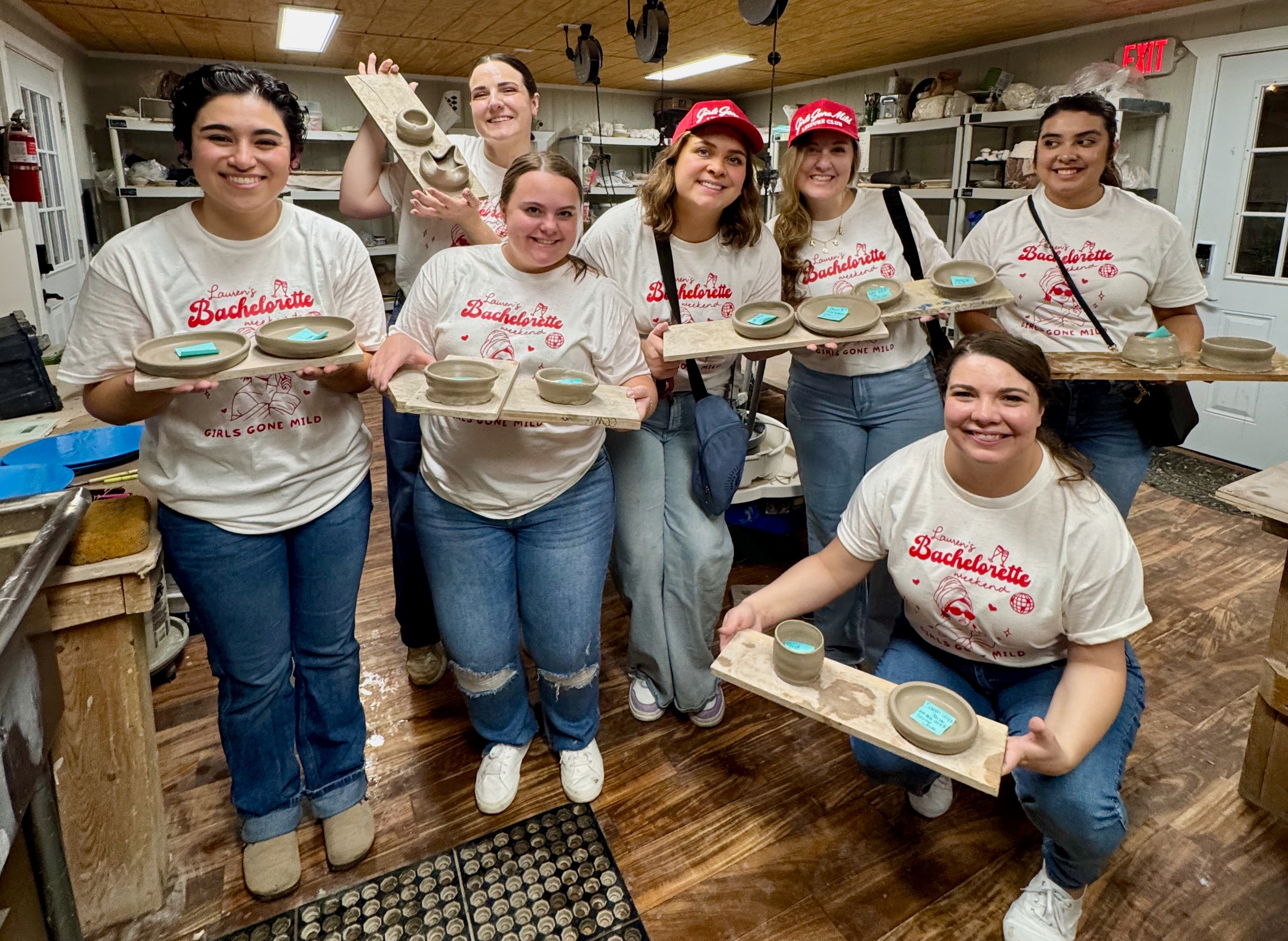 Group of eight young women posing indoors holding pottery pieces on wooden boards, all wearing white t-shirts with red text that says "Laurens Bachelorette Weekend Girls Gone Mild."