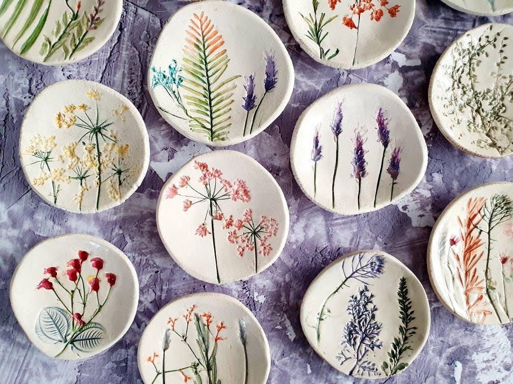 Collection of small ceramic plates with hand-painted floral and botanical designs in various colors, scattered on a textured surface.