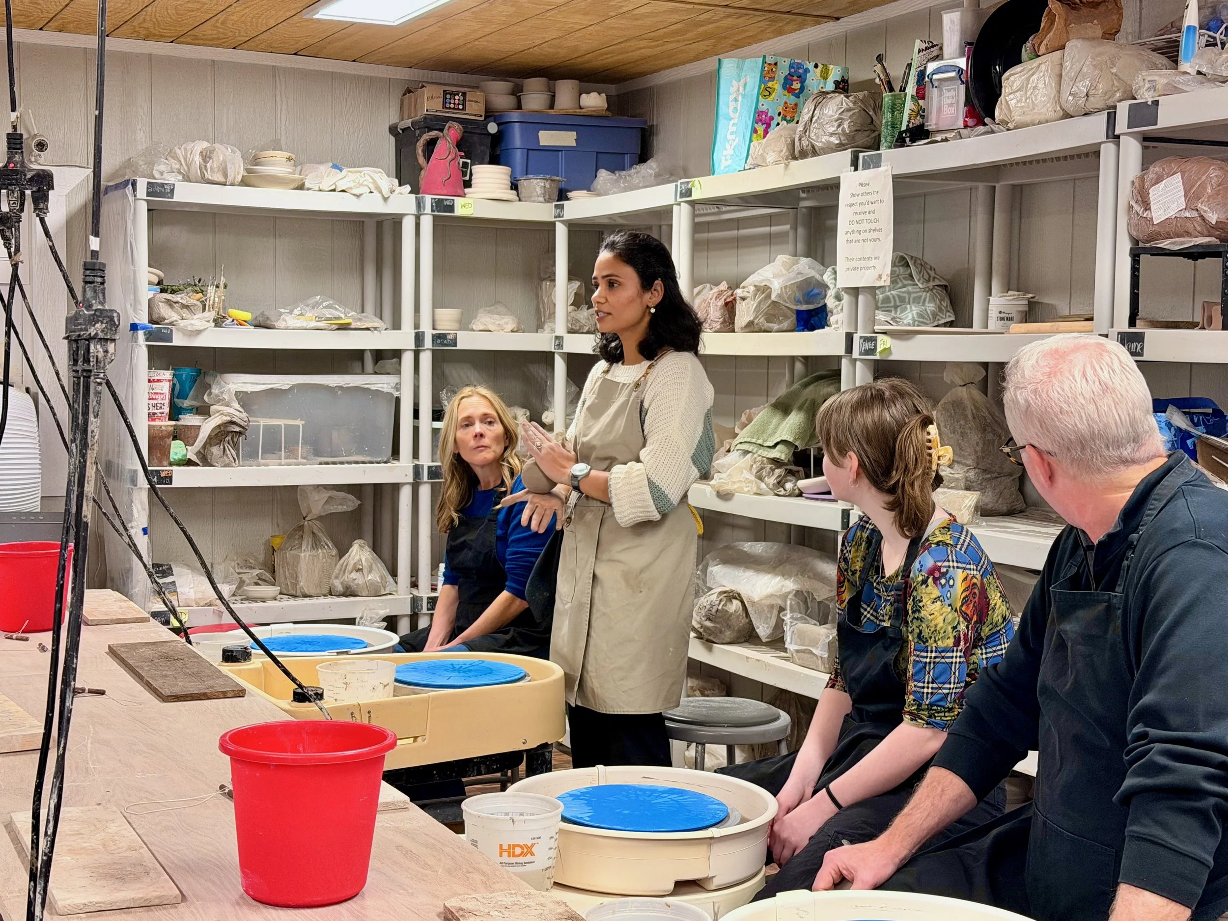 A woman instructing a group in a pottery studio with shelves filled with supplies and clay, some people seated around a table with pottery wheels.