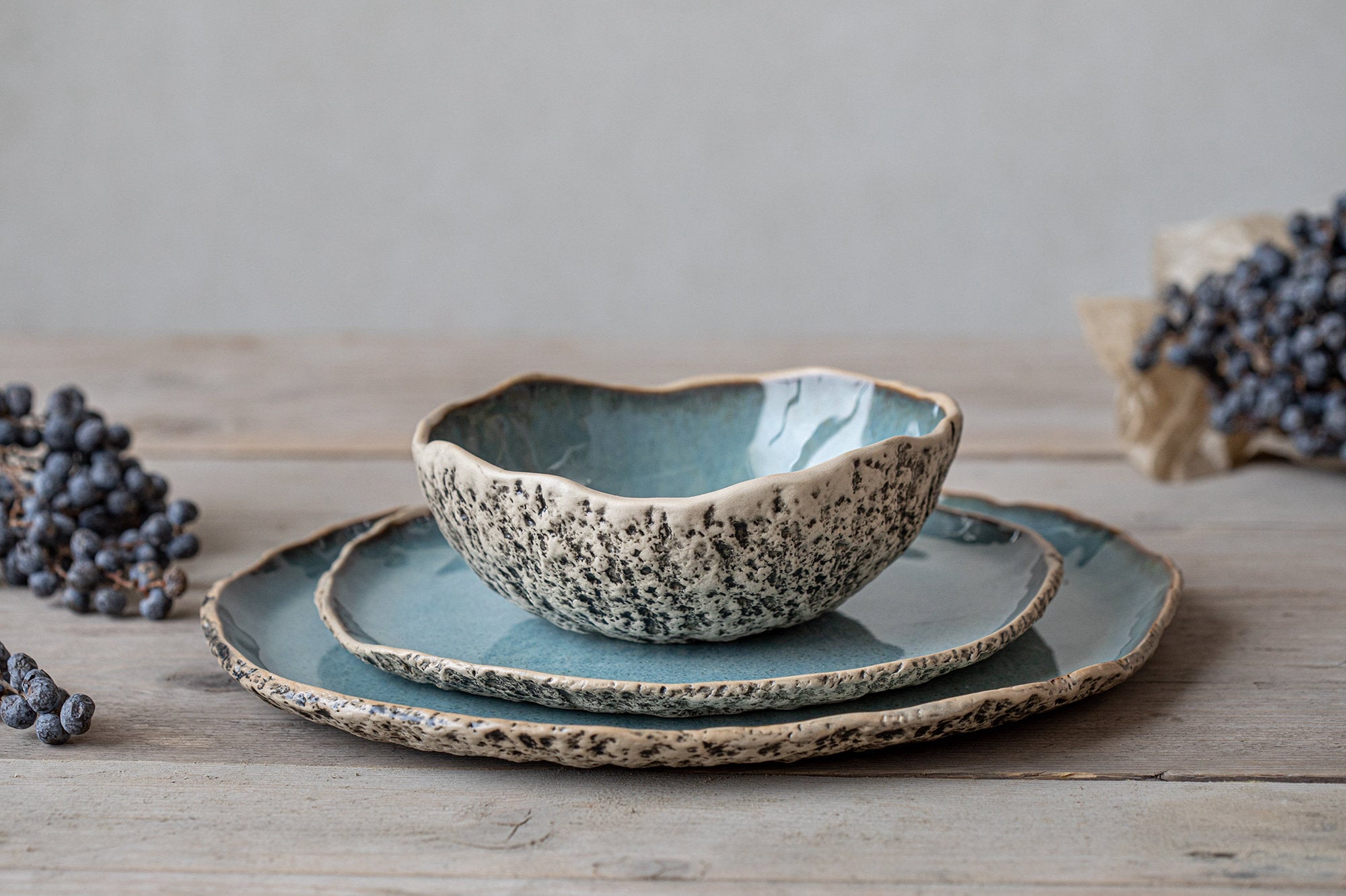Set of three ceramic dishes with a speckled exterior and blue interior, stacked on a light wooden surface, surrounded by bunches of black grapes.