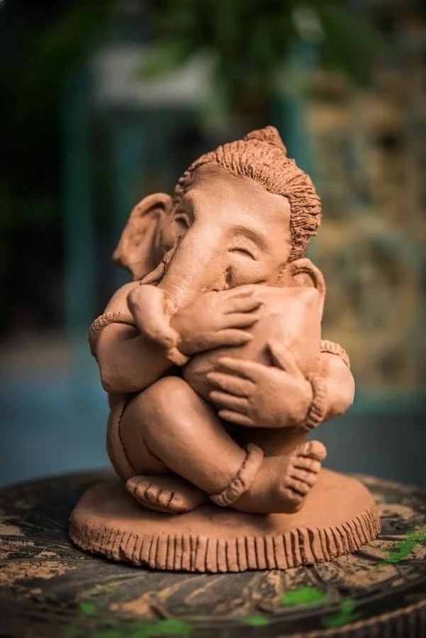 Clay sculpture of a seated character with elephant-like features, holding an object, positioned on a textured surface.