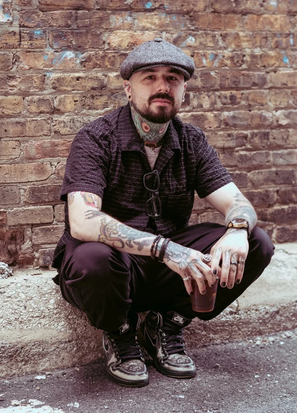 A man with a beard and tattoos, wearing a grey cap, black textured short-sleeve shirt, black pants, and stylish boots, sitting on a ledge in front of a brick wall, holding a drink in one hand.