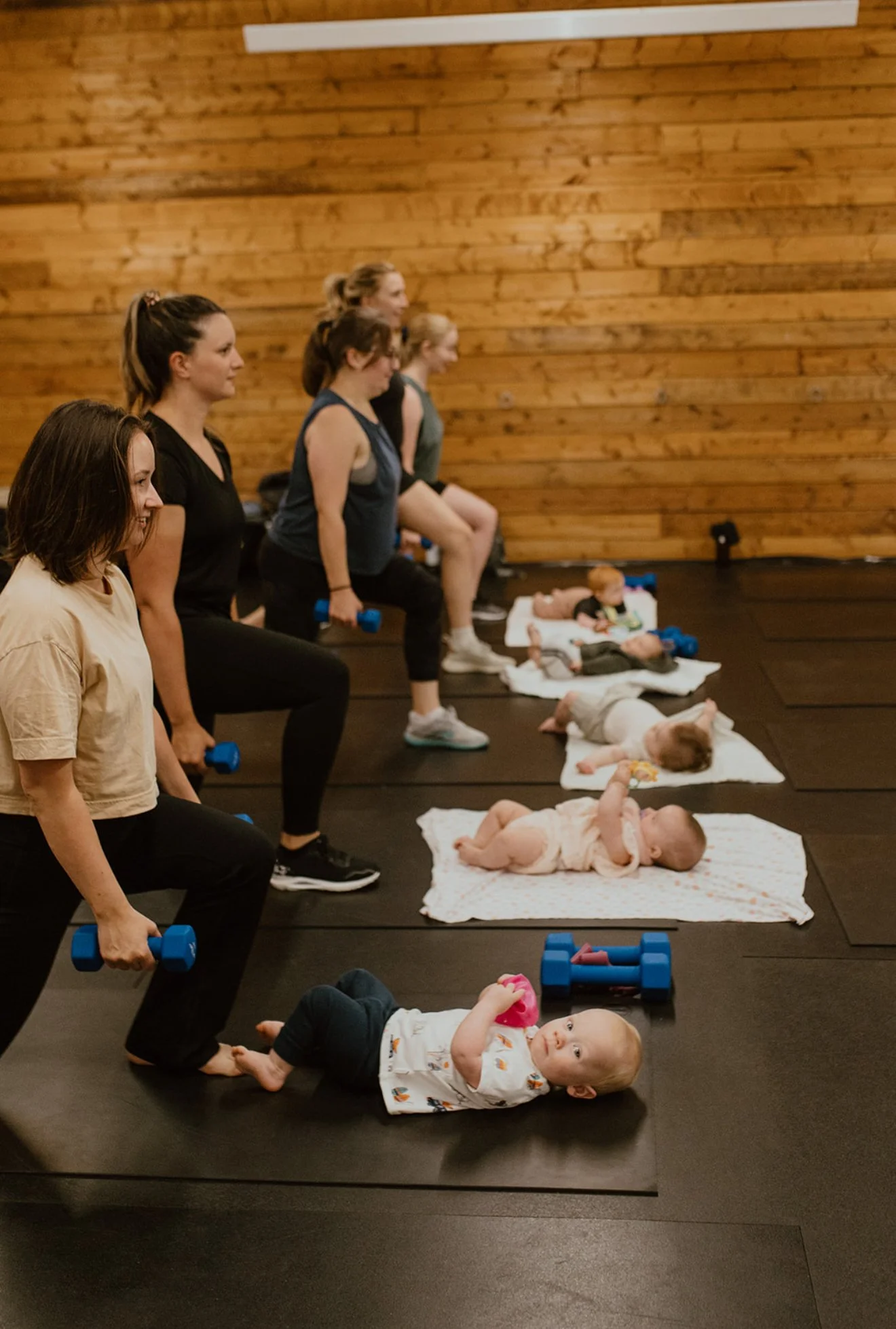Women participate in a fitness class with babies lying on mats in front of them, in a wooden-floored room.