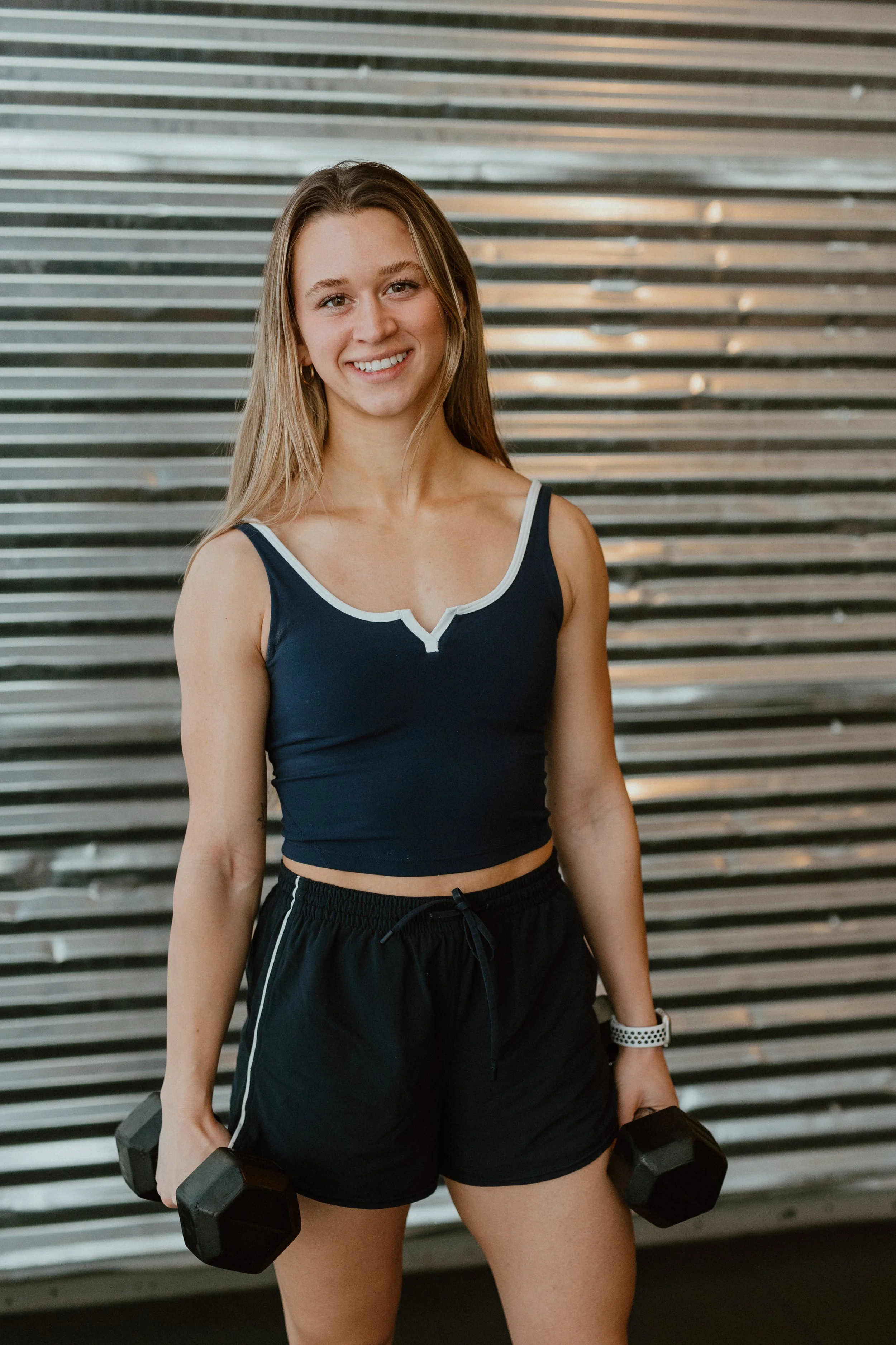 Young woman with long curly brown hair smiling, wearing a black t-shirt and black pants, standing with arms crossed against a metal slat wall background.