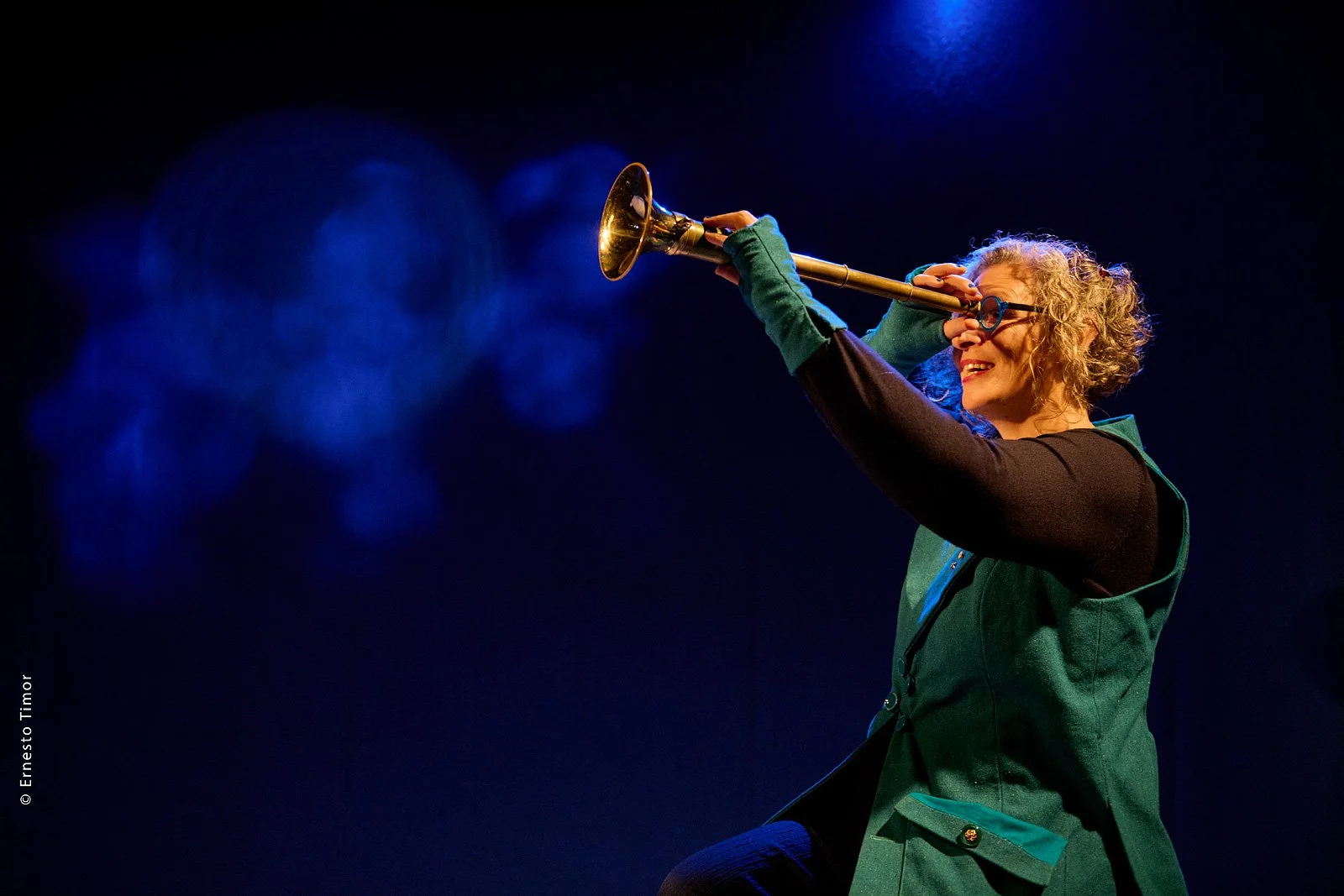 Une femme avec des cheveux bouclés, portant des lunettes et un gilet vert, joue du trombone sur scène avec un fond sombre et des lumières bleues