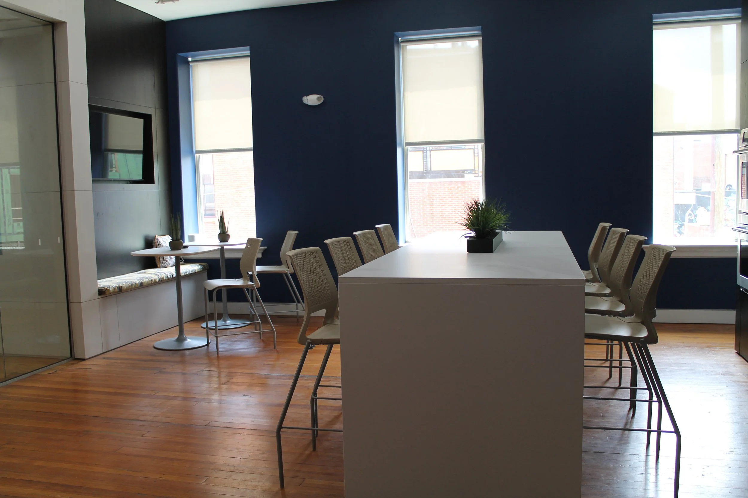 Modern office kitchen with a large white table, beige chairs, blue accent walls, a built-in window seat with cushions, and small potted plants on the tables.