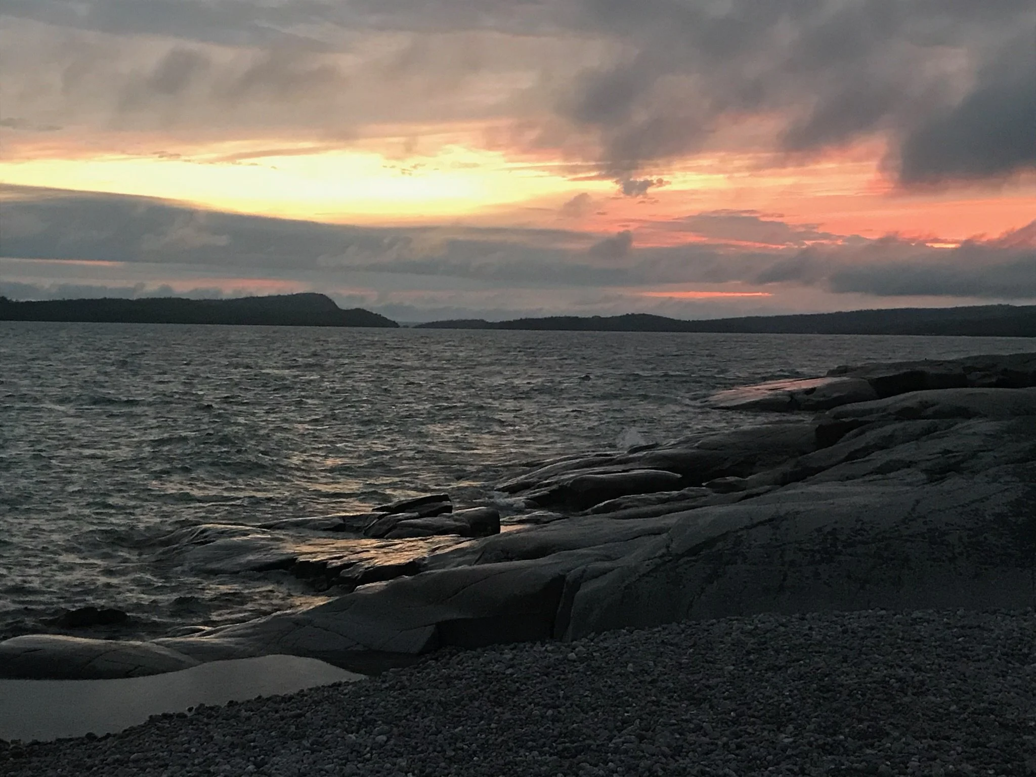 A rocky shoreline under a cloudy sky at sunset, with dark clouds and pinkish-orange hues on the horizon, calm water, and distant landmasses.
