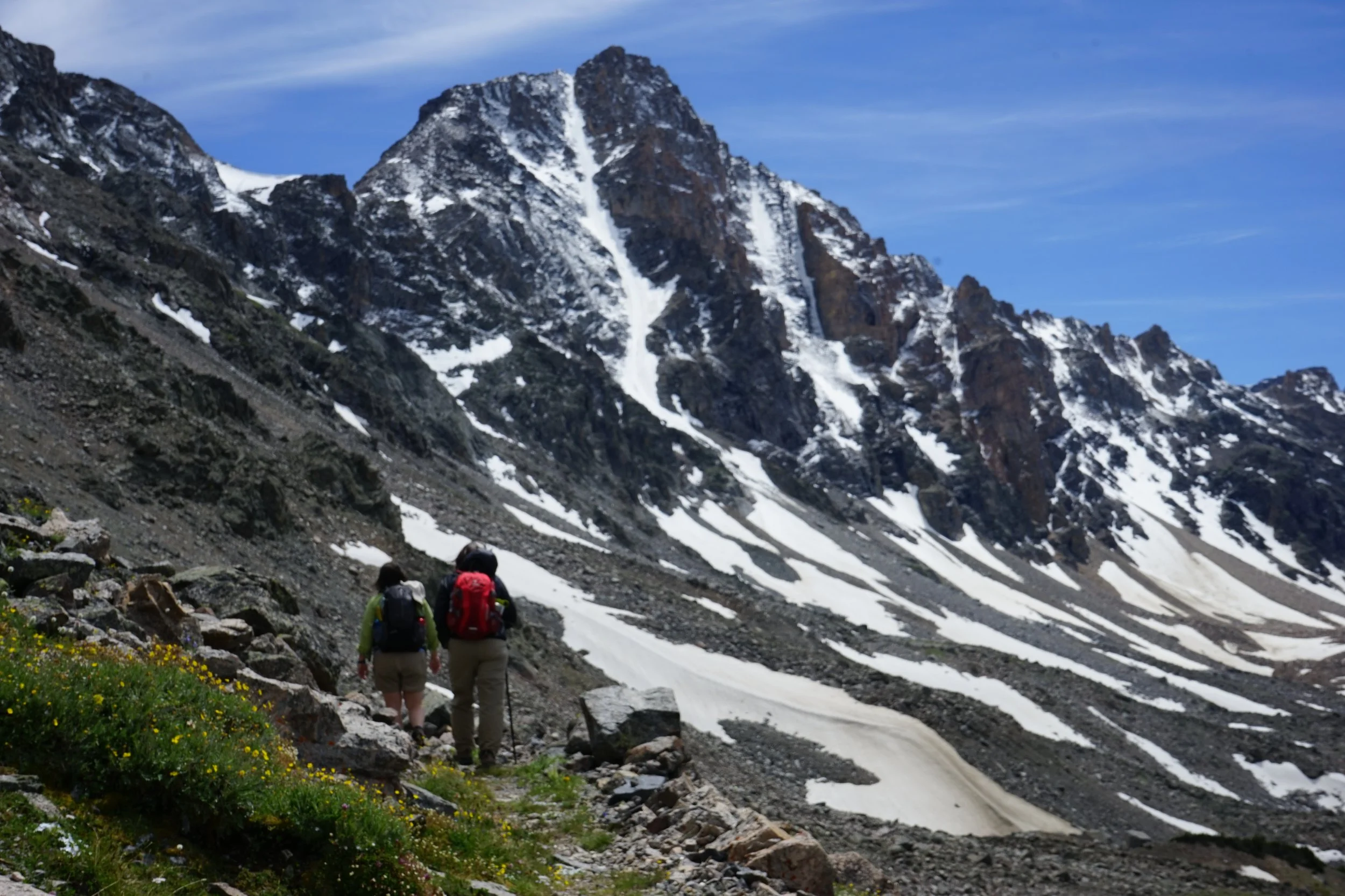 Two hikers with backpacks walking along a mountain trail with snow patches on a rocky slope under a blue sky.