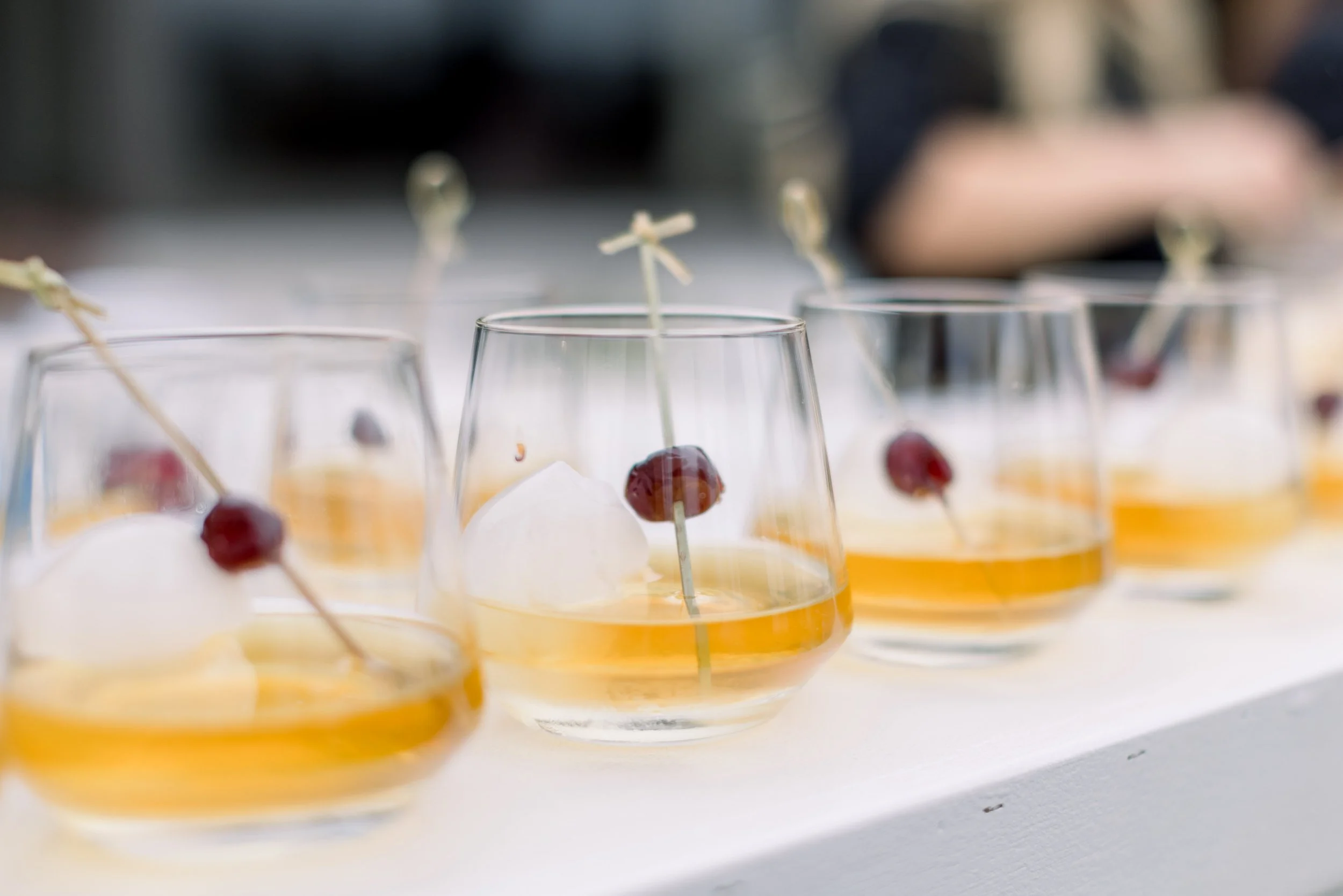 Close-up of small clear glasses containing a yellow-orange liquid, garnished with a maraschino cherry and topped with white foam, lined up on a white surface.