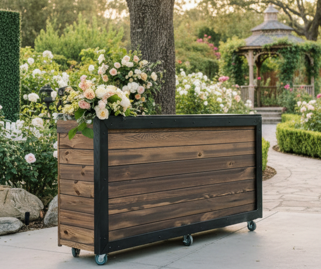 A wooden planter box on wheels filled with pink and white flowers, situated on a paved garden path with a lush garden and gazebo in the background.