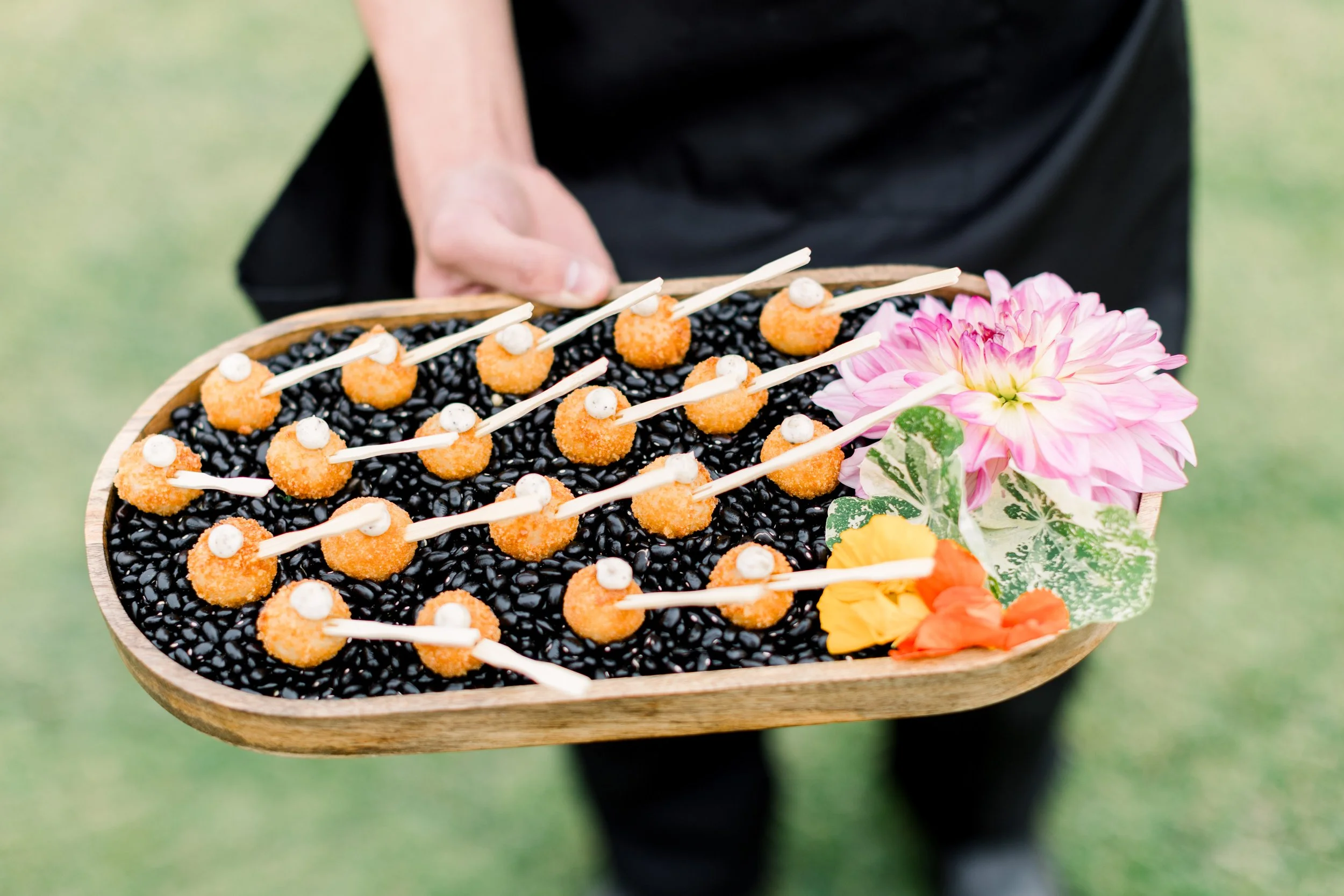 Person holding a wooden tray of fried appetizers topped with white sauce, surrounded by pink and orange flowers, against a blurred green outdoor background.