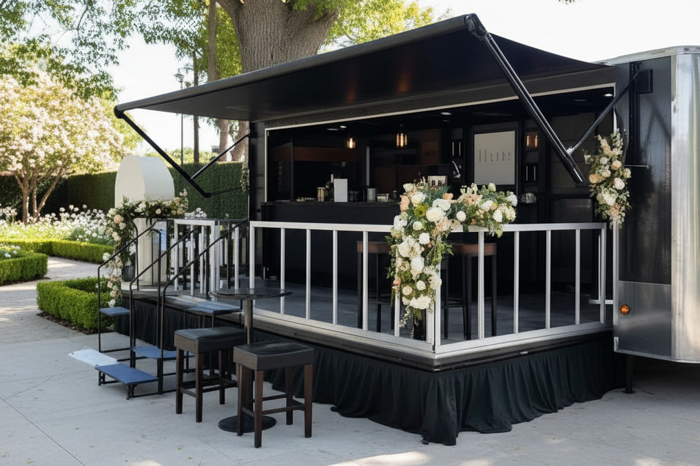 A black mobile bar trailer decorated with white and cream floral arrangements, set outdoors on a paved area with trees and hedges in the background.