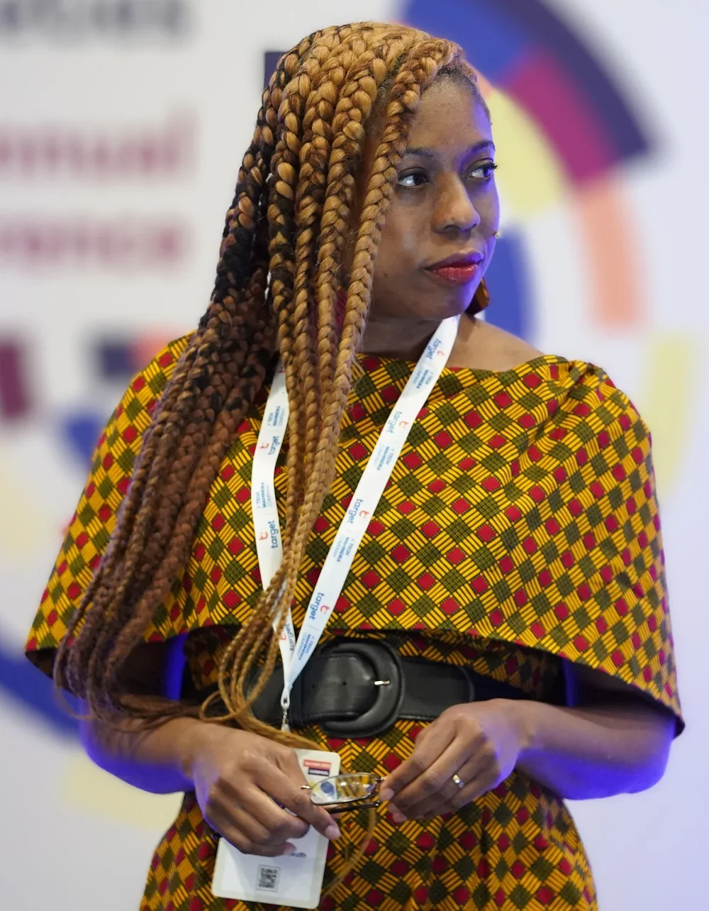 A woman with long, braided hair wearing a colorful, patterned dress and a conference badge around her neck.