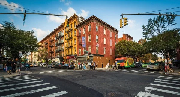 City street intersection with pedestrians, cars, a colorful bus, and historic brick buildings, under a partly cloudy sky.
