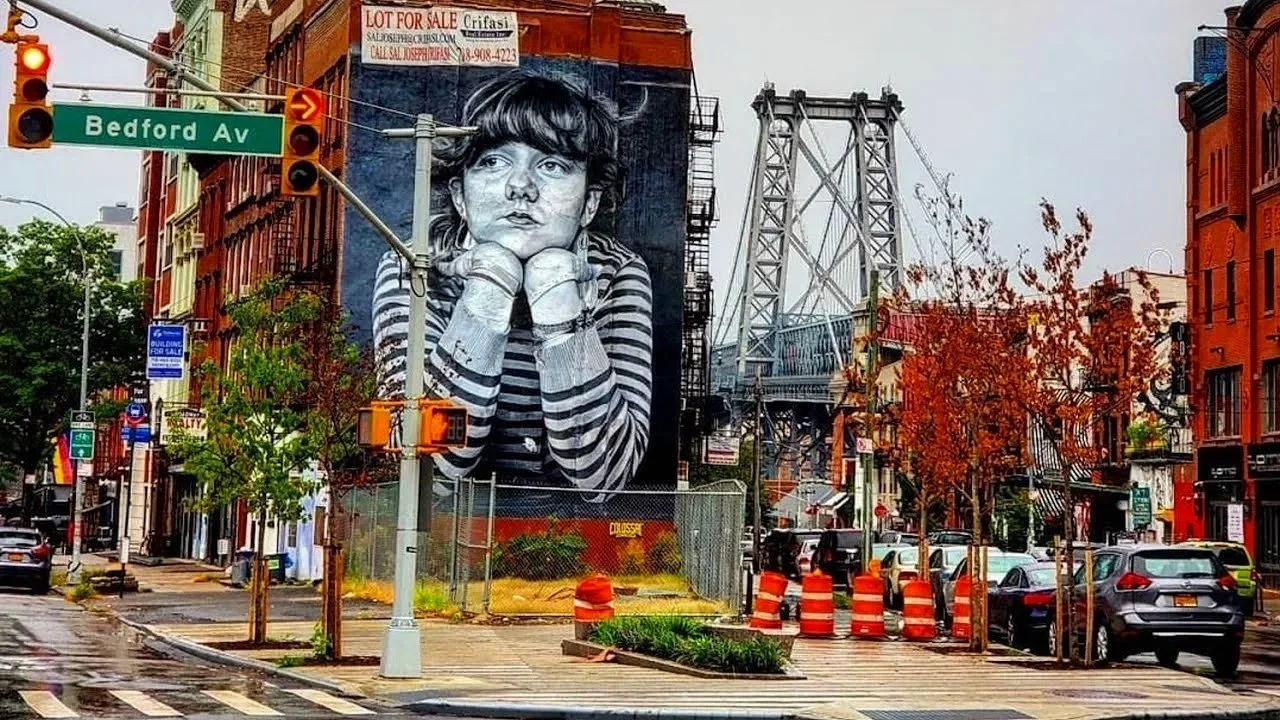 Street scene featuring a large mural of a young girl in black and white, resting her chin on her hands, on the side of a building. The mural is situated at an intersection with traffic lights and signs, including one that reads 'Bedford Ave' and another that says 'LOT FOR SALE.' The scene includes a bridge in the background and a row of parked cars along the street, with trees and orange traffic barrels in the foreground.