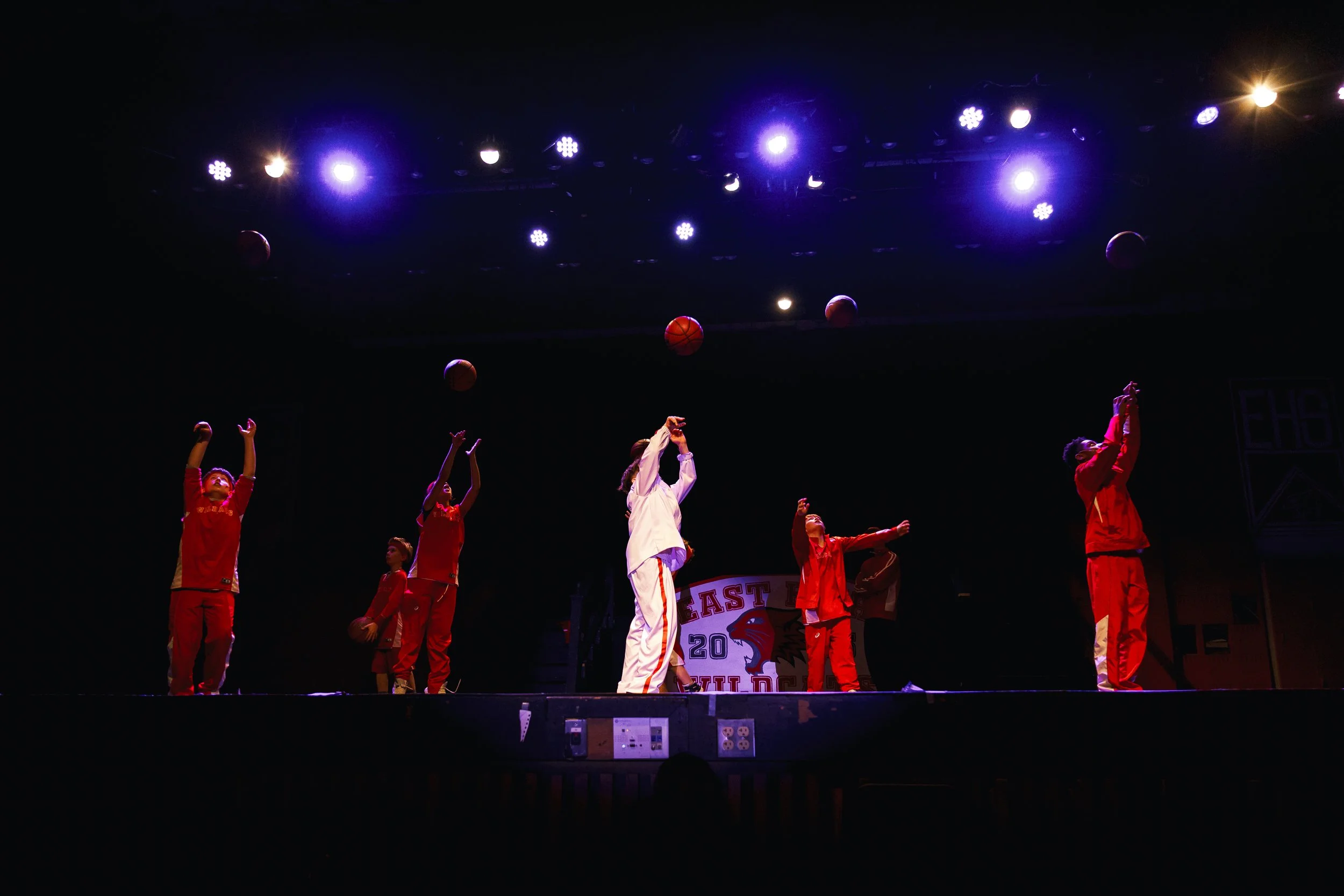 Young basketball players practice shooting on stage with purple lights overhead at a school event.