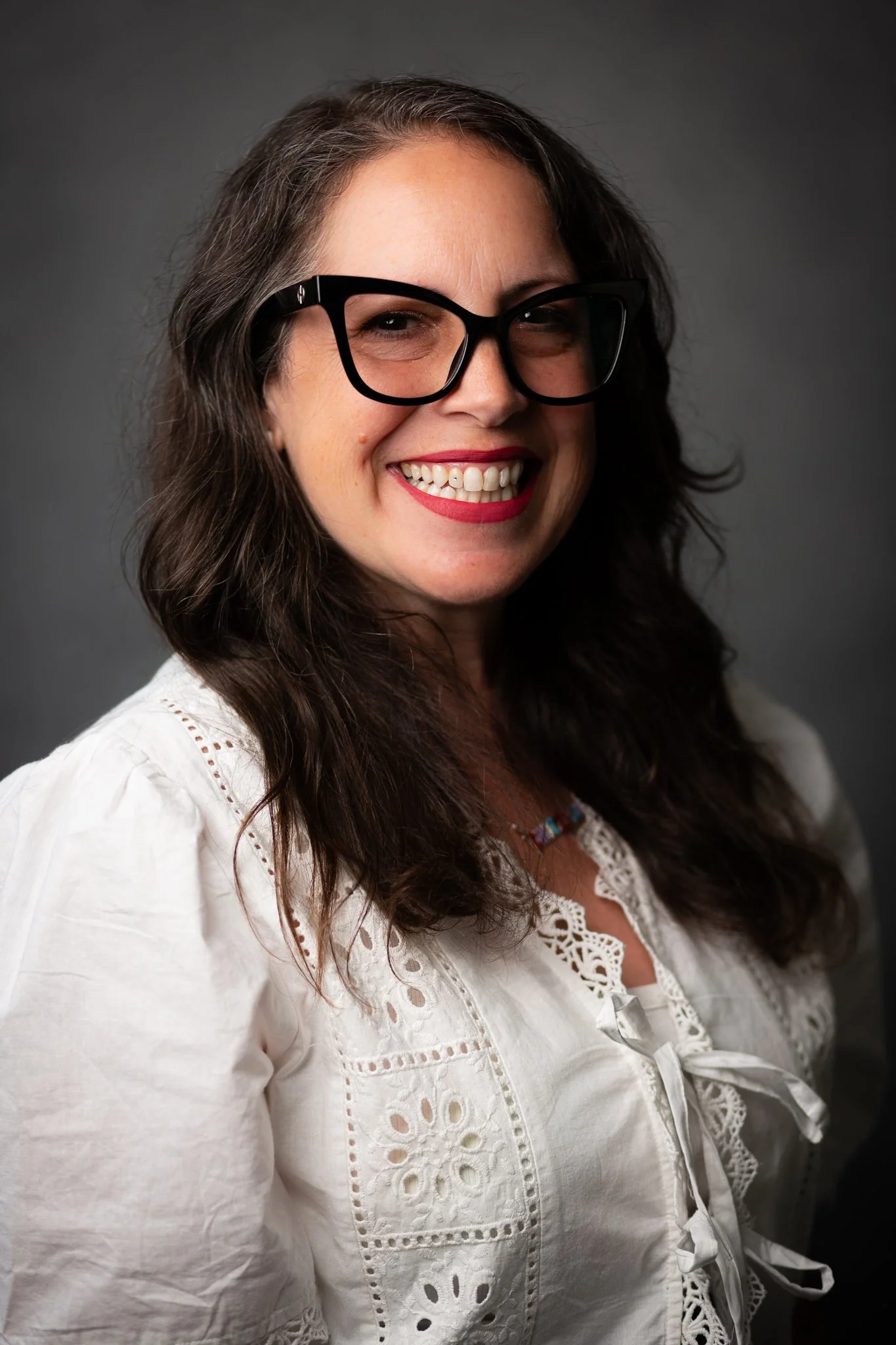 A woman with dark wavy hair wearing glasses and a white embroidered blouse, smiling against a gray background.