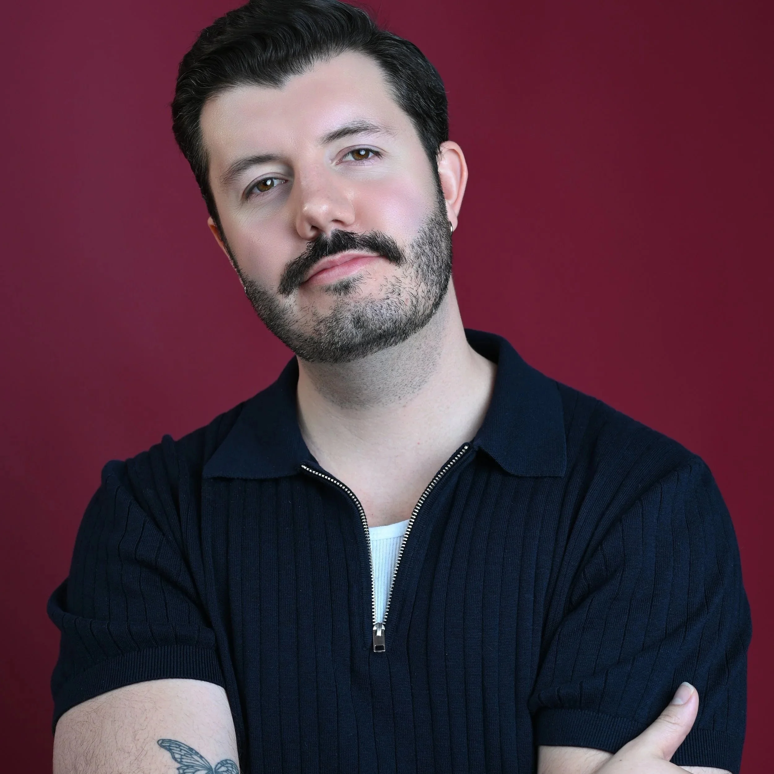 A man with dark hair, a beard, wearing a black collared shirt with a zipper, standing against a solid maroon background.