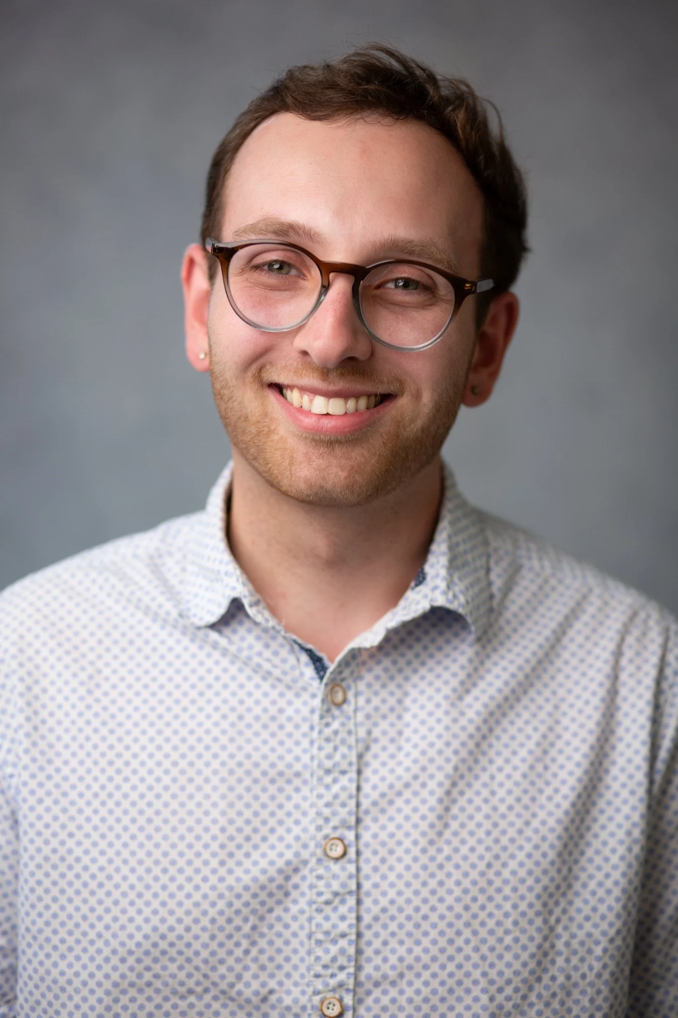 A young man with glasses and short hair, smiling, standing in front of a brick wall, wearing a fleece jacket over a sweater.