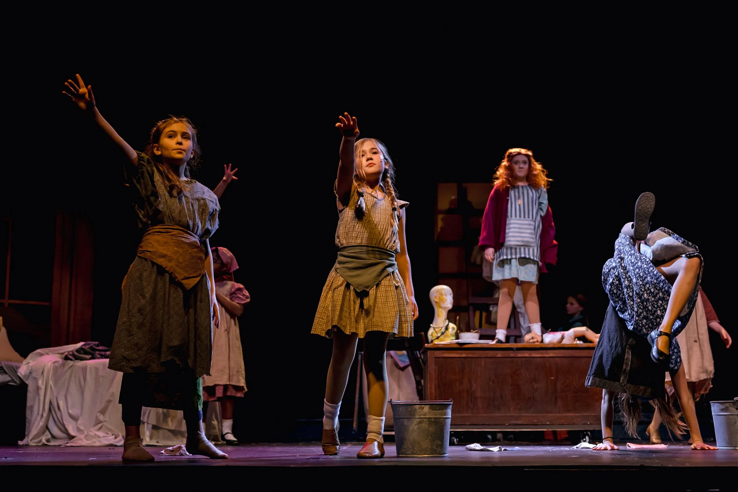 Children performing a theatrical play on stage, with some acting and others in various poses, set against a dark background with stage props and costumes.