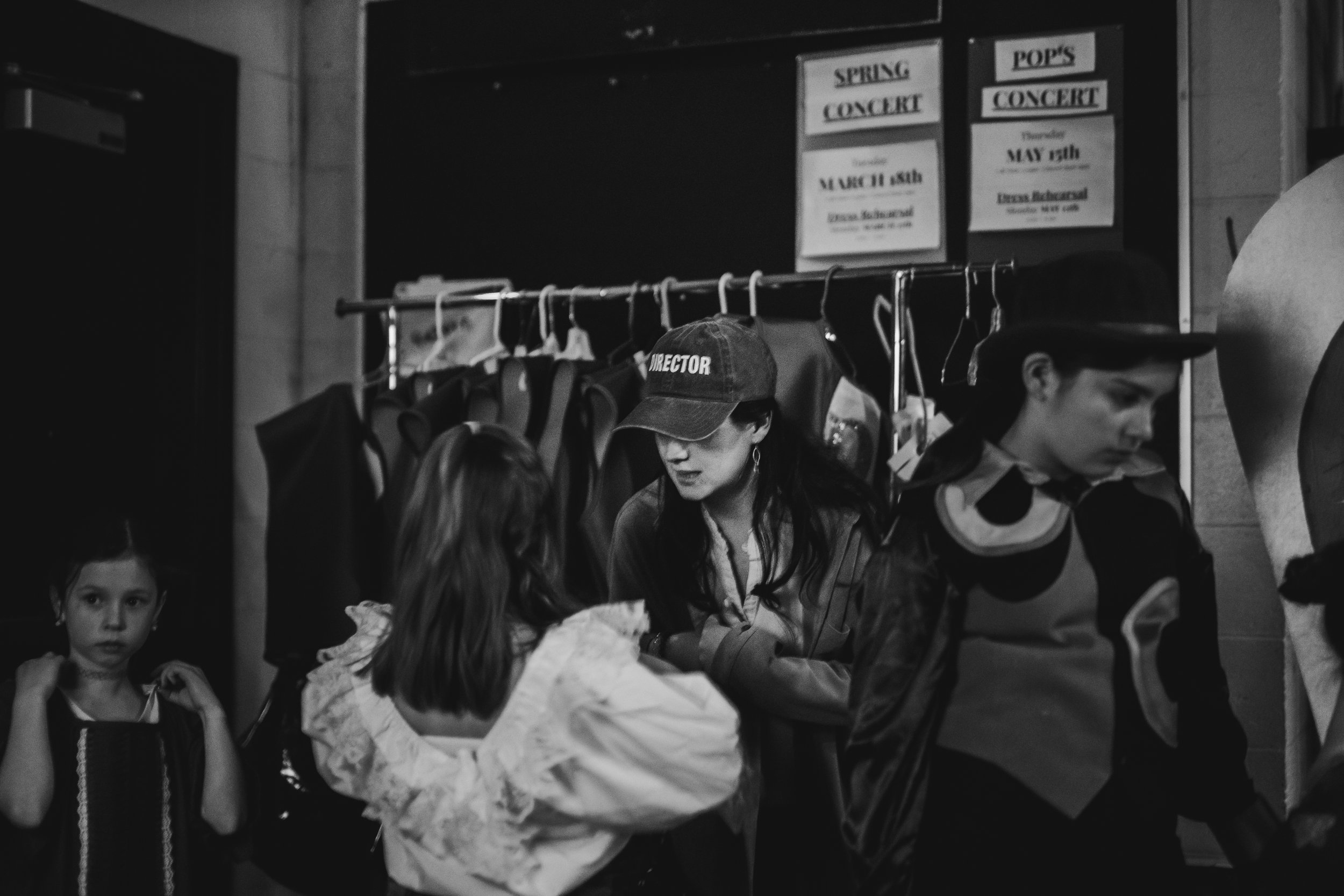 A woman with a cap labeled 'DIRECTOR' talking to two young girls in a backstage area with costumes hanging on a rack and posters on the wall for spring and pop concert events.